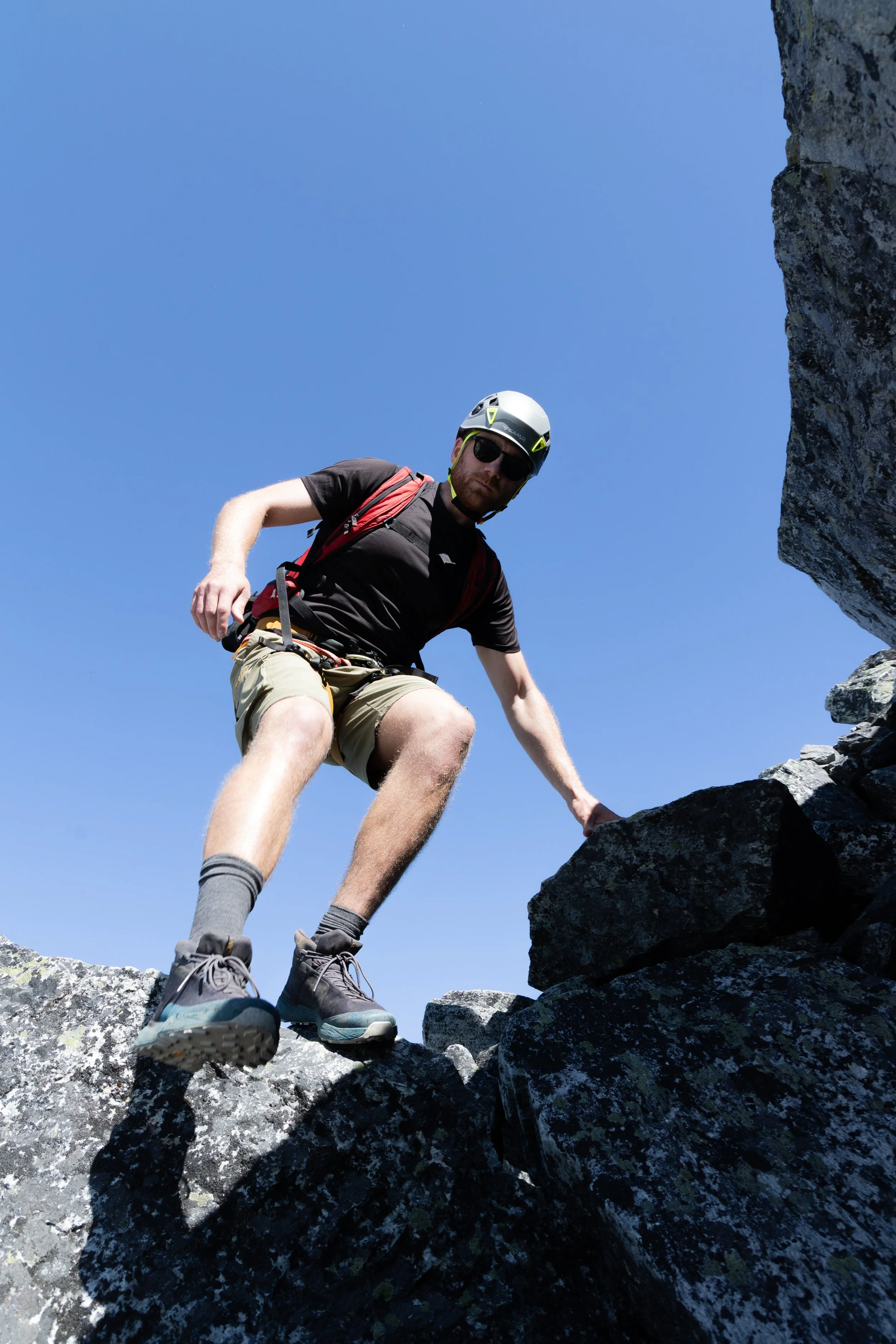 Man wearing a helmet, sunglasses, and a black t-shirt climbing over rocks on a mountain trail on a clear day.