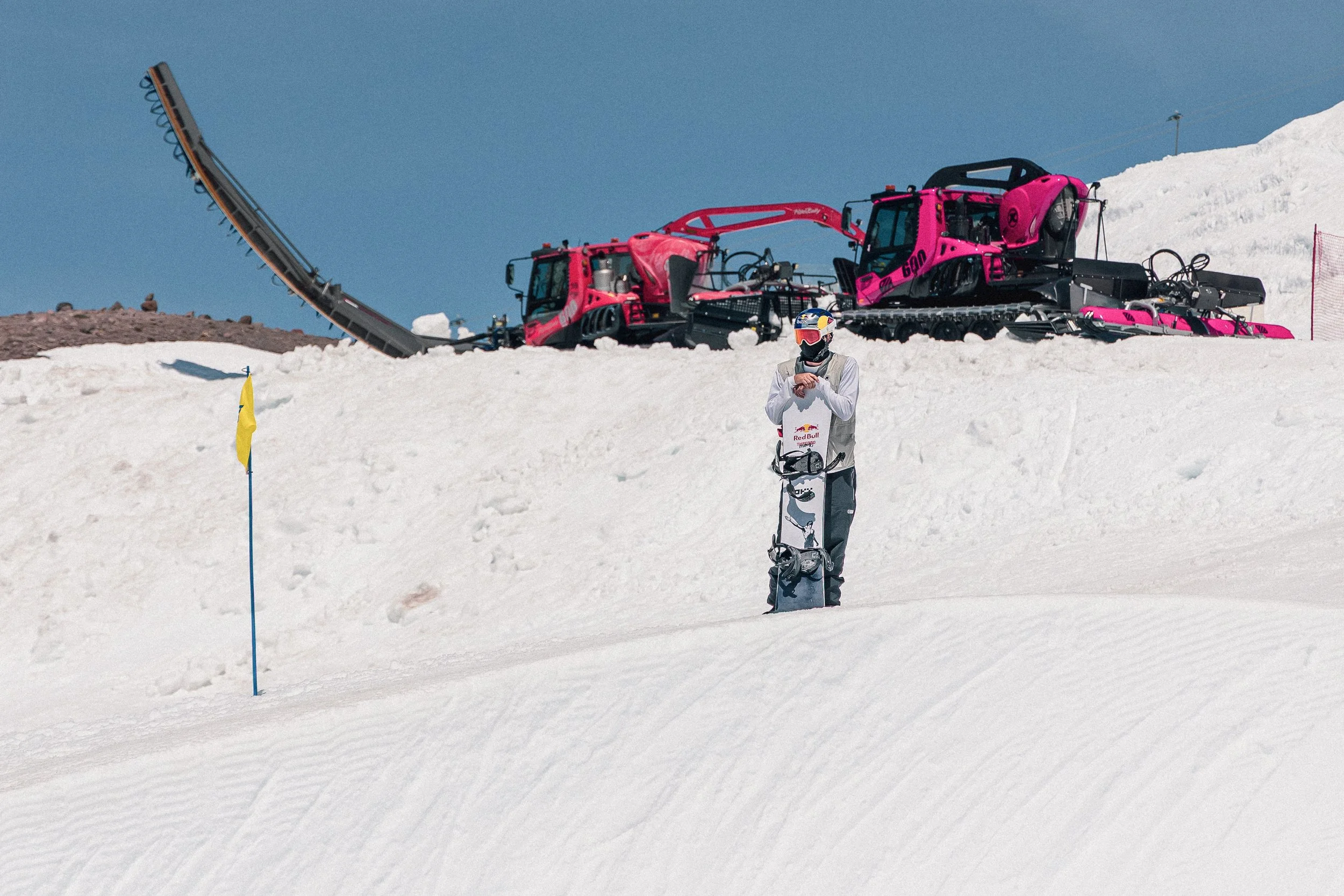 A person dressed in snowboarding gear standing on a snow-covered slope with a snowboard, near yellow flag, with large pink snow grooming machines in the background under a clear blue sky.
