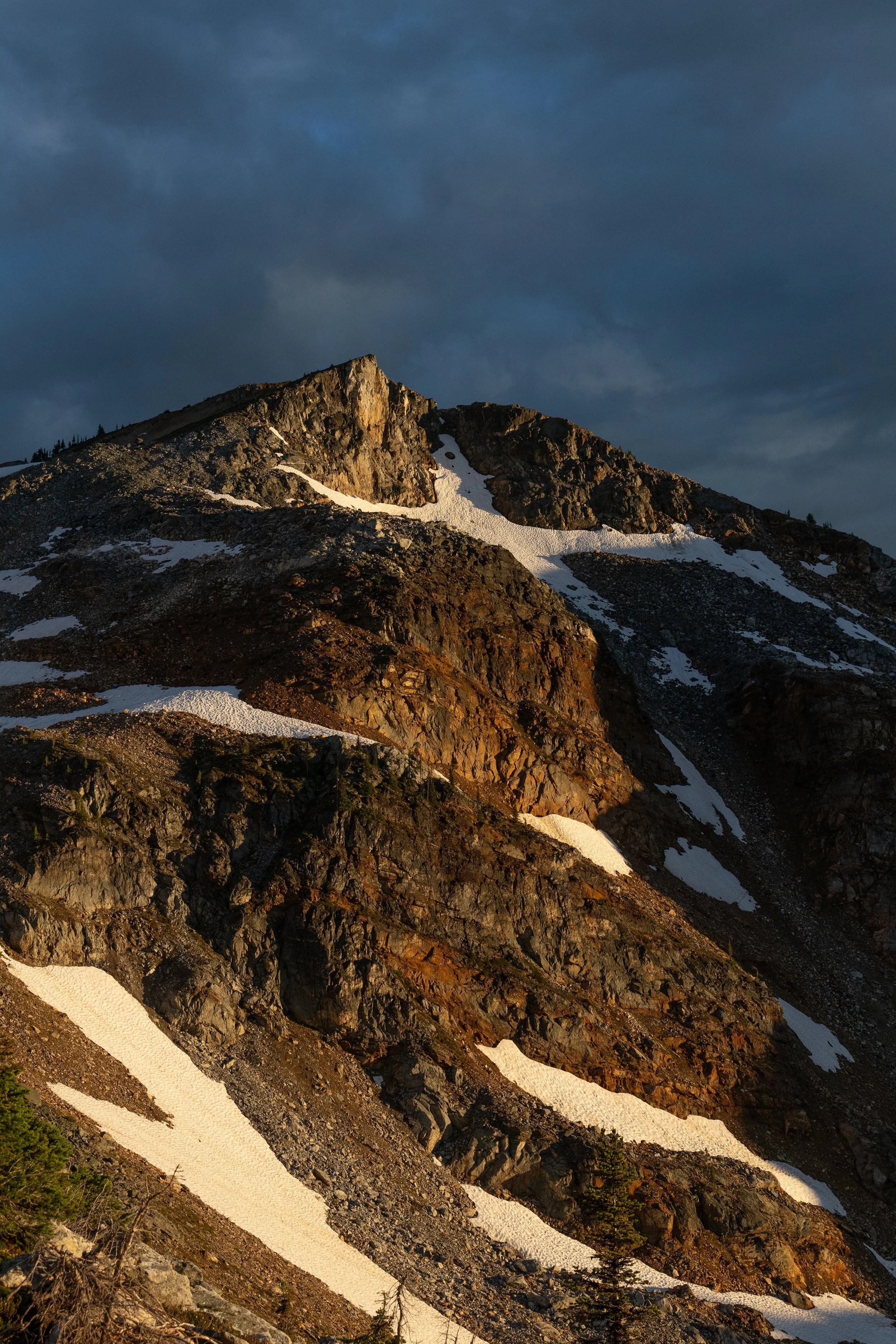 A rocky mountain peak during sunset with patches of snow and scattered trees in the foreground under a cloudy sky.