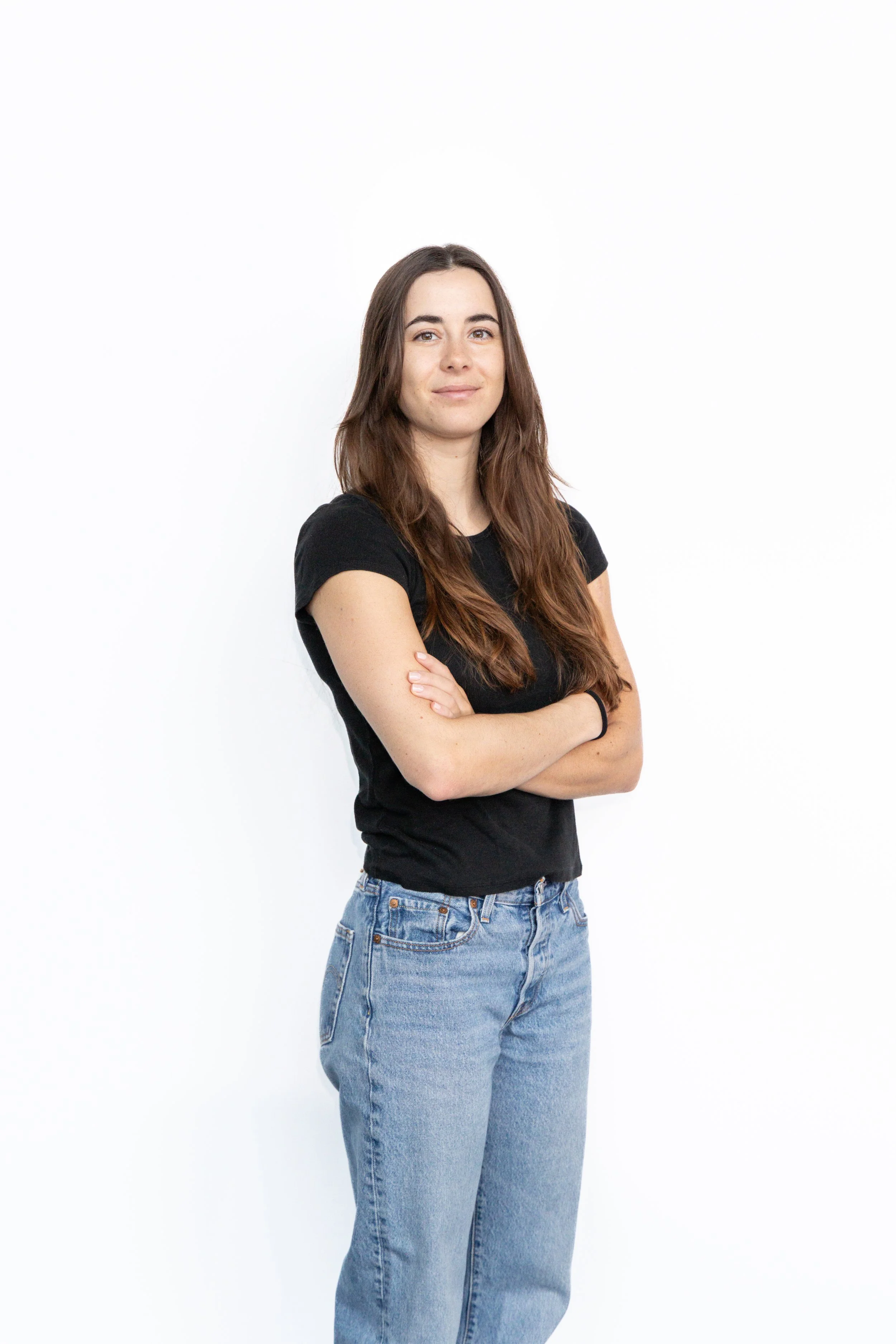 A young woman with long brown hair wearing a black T-shirt and high-waisted blue jeans, standing with arms crossed against a plain white background.