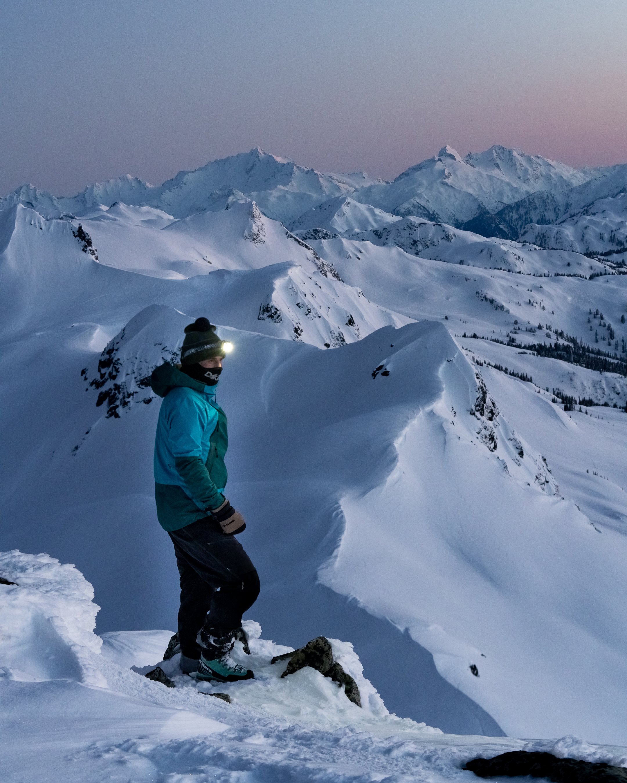 A person dressed in winter gear, including a blue jacket, black pants, and a hat with a headlamp, standing on snowy mountain terrain at dusk or dawn with snow-covered peaks in the background.