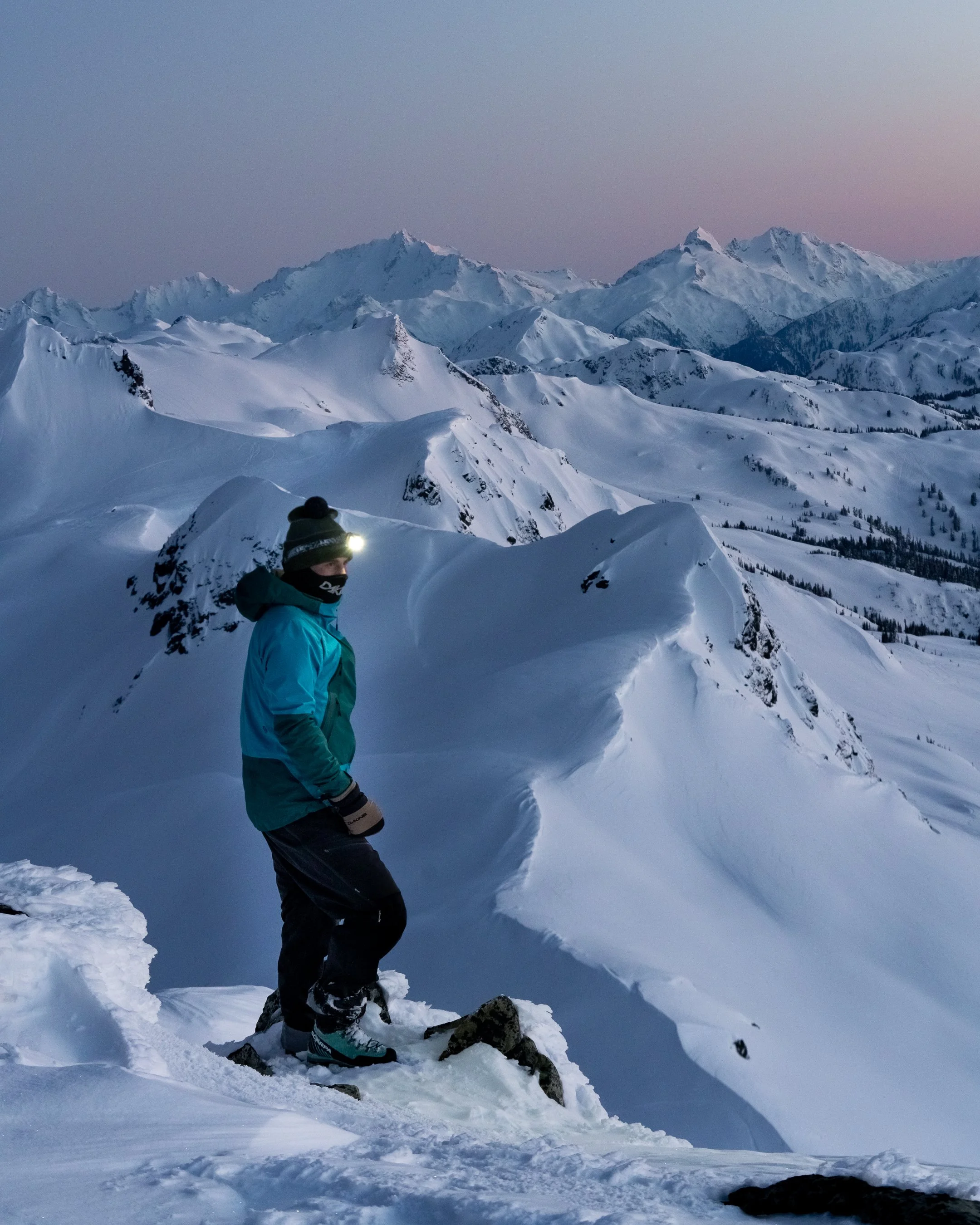 A person dressed in winter gear, including a blue jacket, black pants, and a hat with a headlamp, standing on snowy mountain terrain at dusk or dawn with snow-covered peaks in the background.