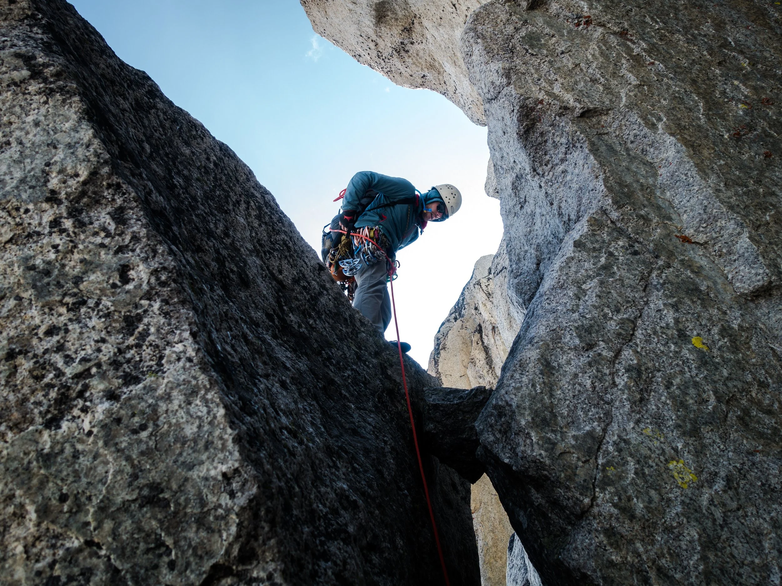 A person wearing climbing gear, including a helmet and harness, climbing inside a narrow rocky crevice with a clear sky visible above.