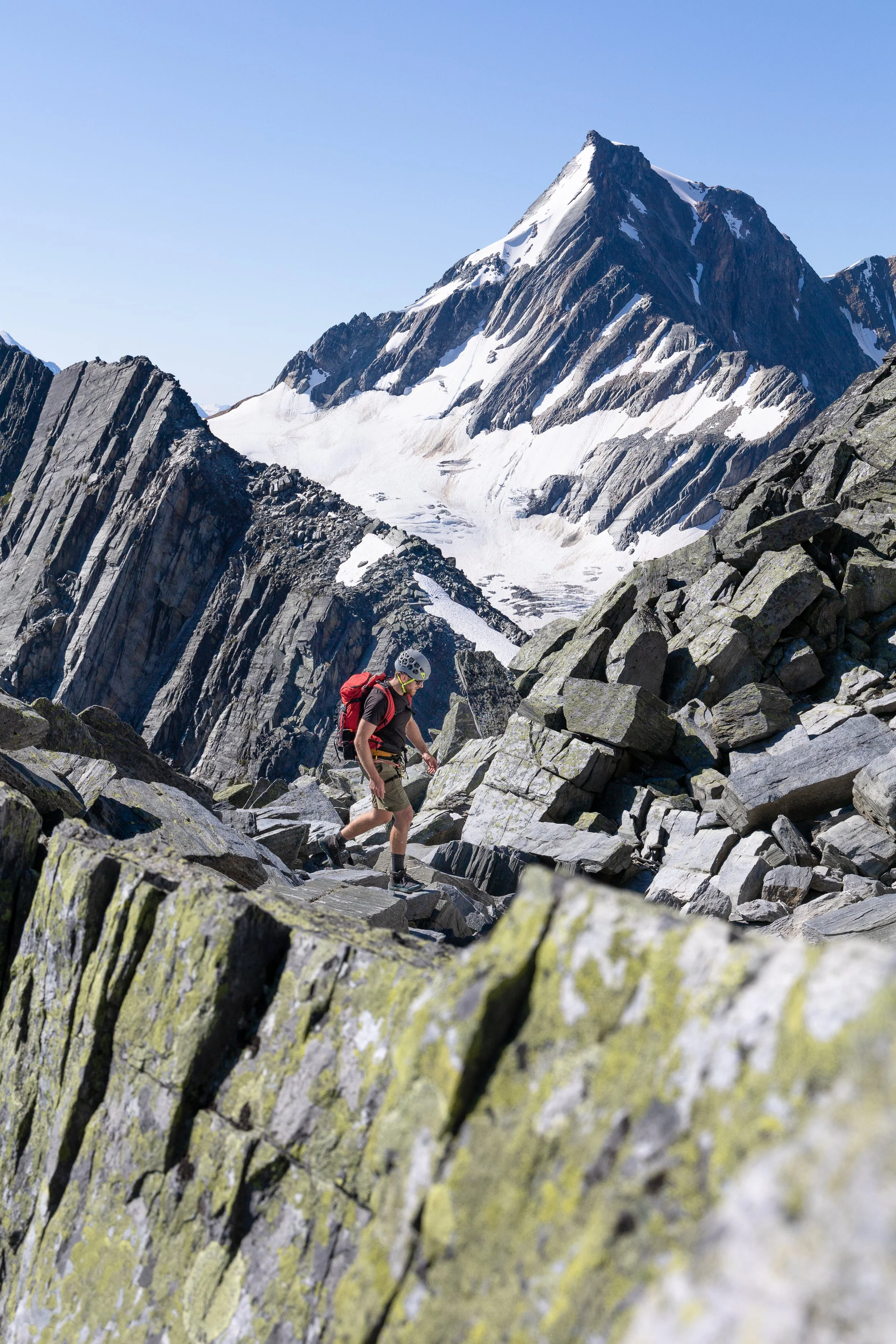 A hiker with a red backpack and helmet trekking through rocky mountain terrain with snow-capped peaks in the background under a clear blue sky.