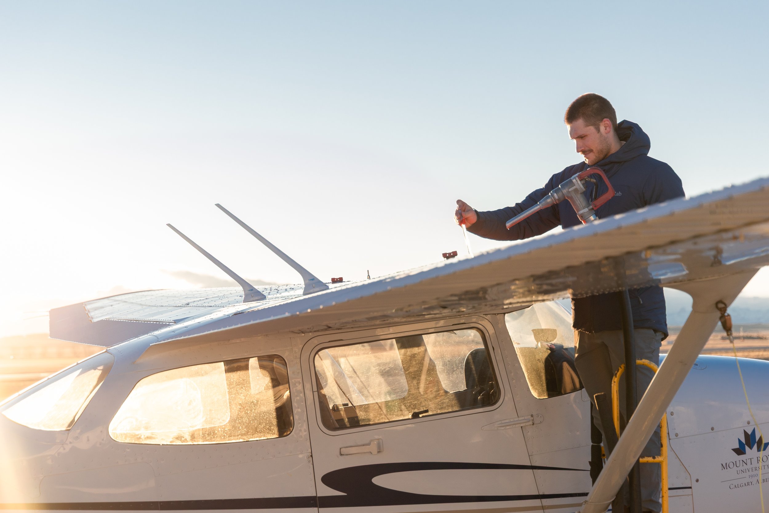 A man in a dark jacket standing on the wing of a small airplane, handling maintenance tools during sunset.