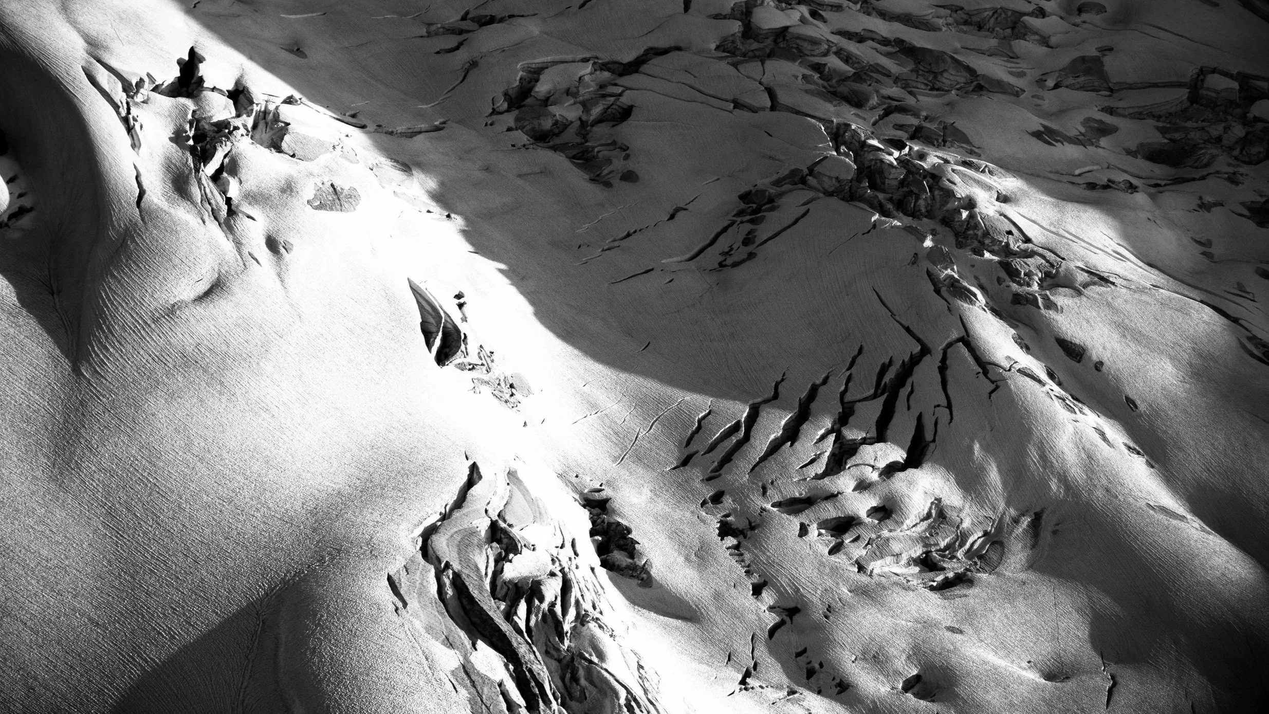 Black and white aerial view of snow-covered mountain terrain with ridges, cracks, and rocky outcroppings.