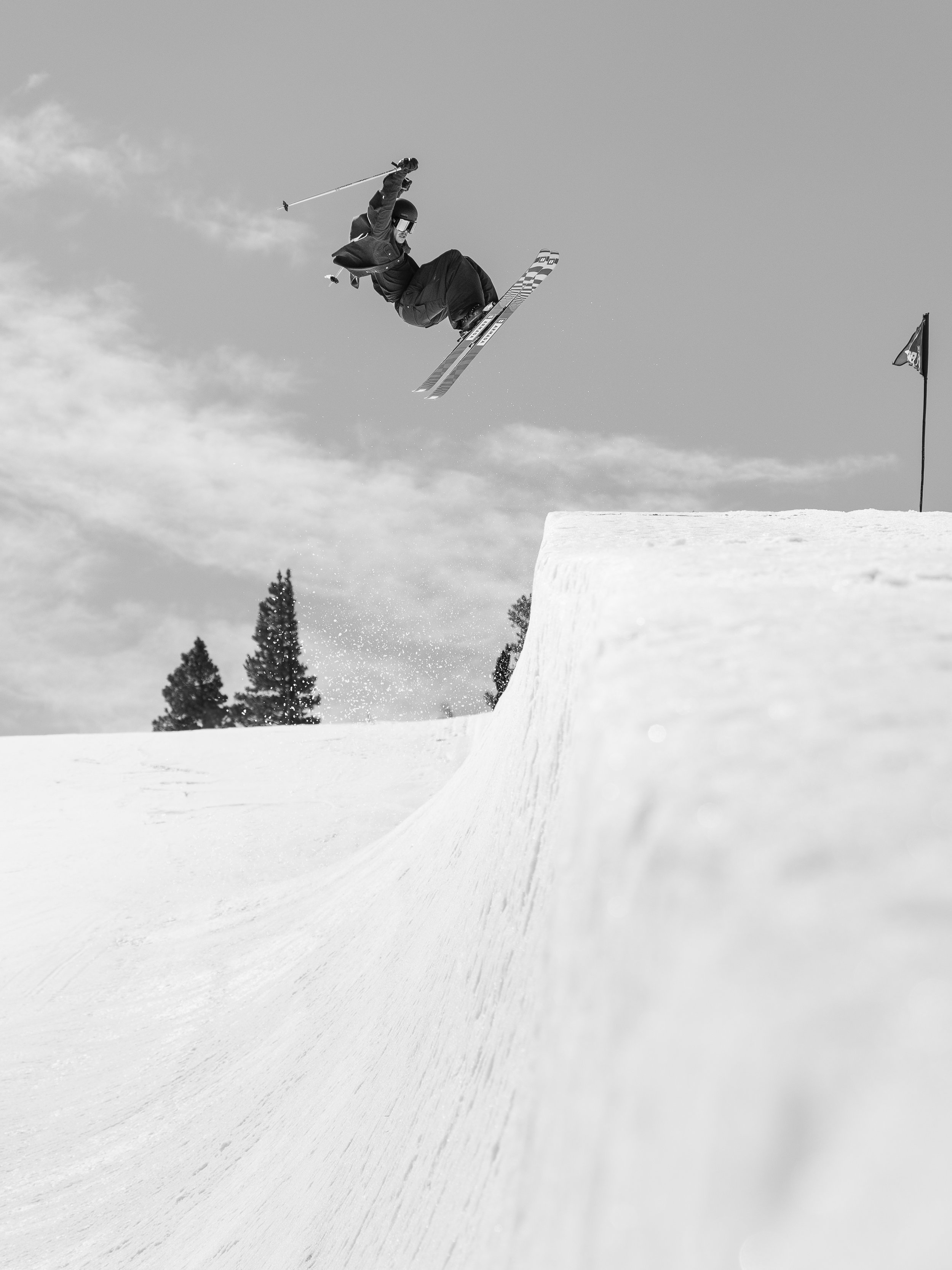 A snowboarder in mid-air performing a jump at a snowy ski resort, with trees and a flag in the background.