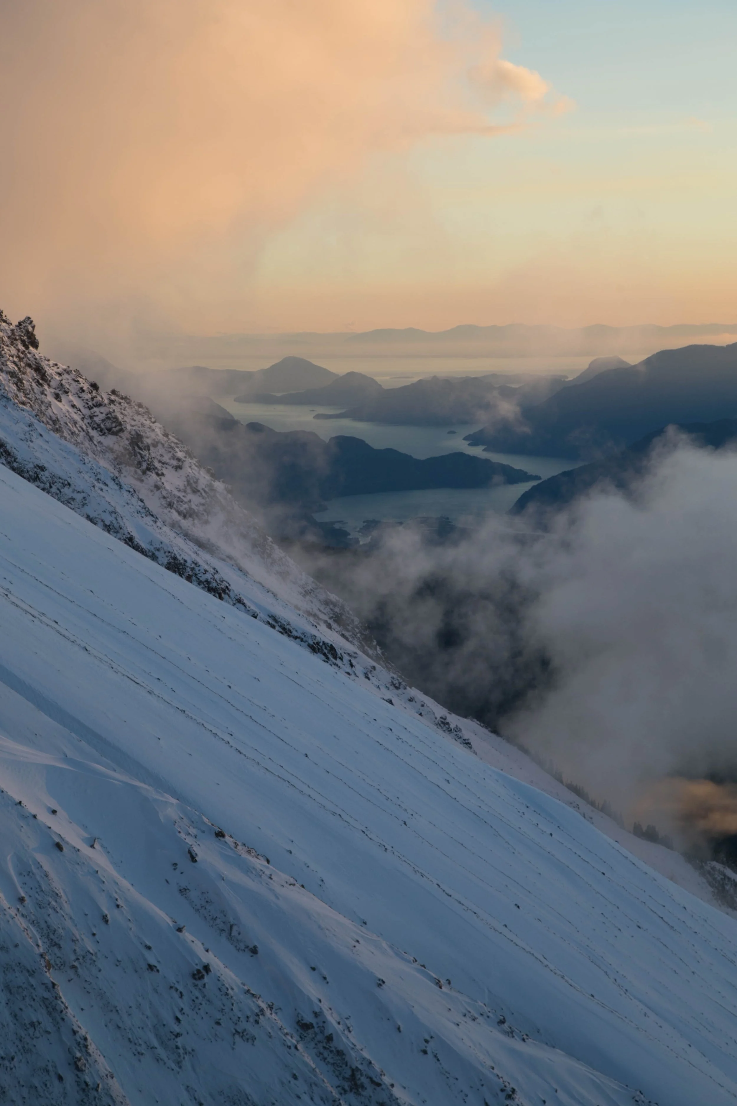 Snow-covered mountain slope with ski tracks, overlooking a mountain lake and distant mountain range at sunset, with clouds and mist.