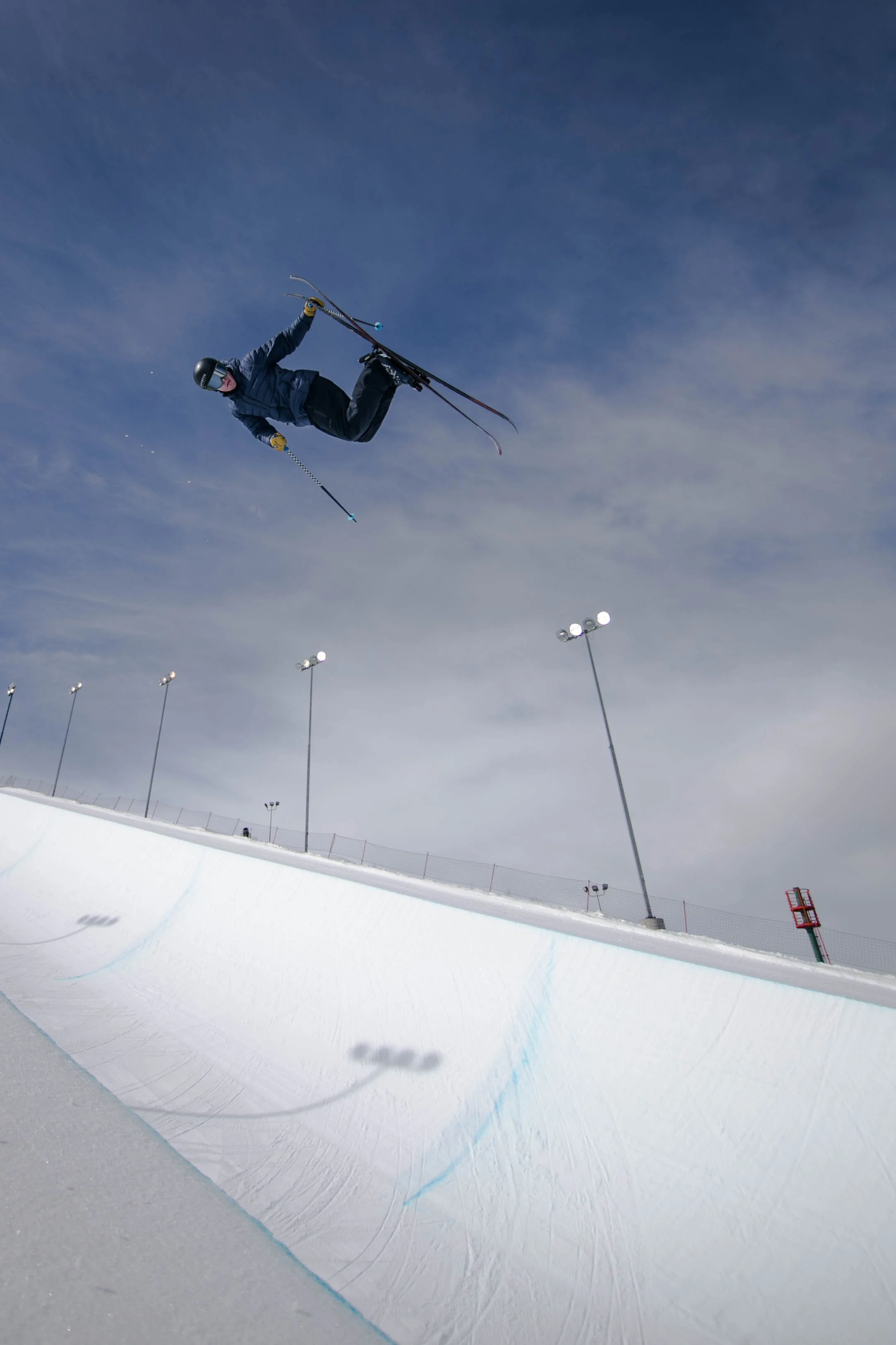 A skier performing a jump in mid-air at a snow-covered ski park, with a cloudy sky and tall light poles in the background.