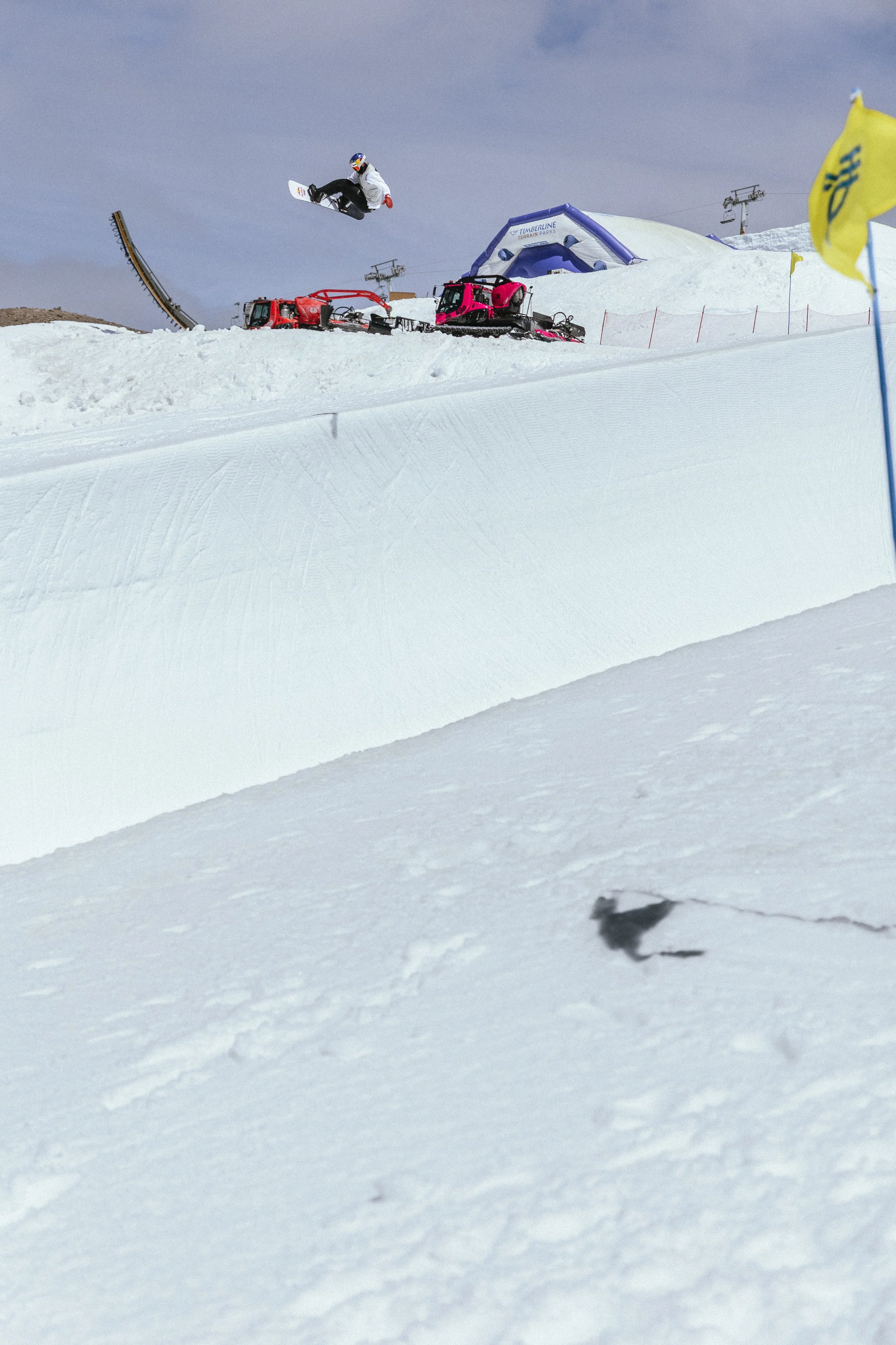 A snowboarder in mid-air performing a jump on a snowy slope at a ski resort. There are snow machines and ski lift infrastructure in the background, along with blue skies and a few flags on the side of the slope.