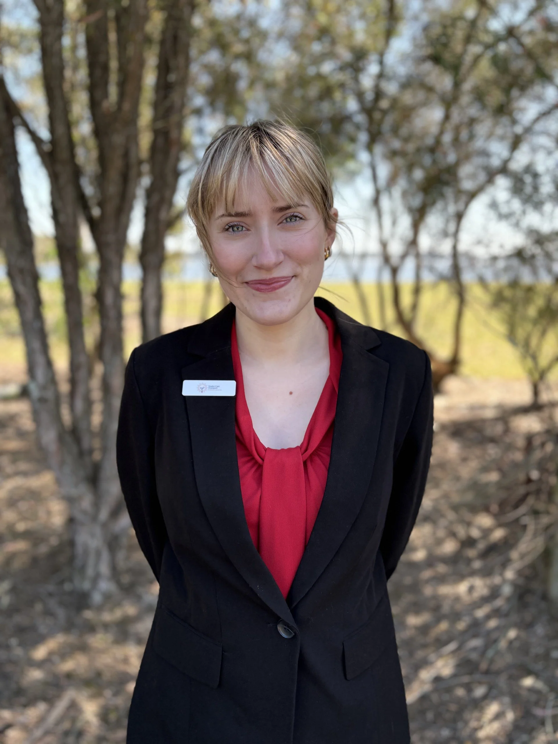 A woman in a black blazer and red blouse standing outdoors with trees in the background, smiling at the camera.