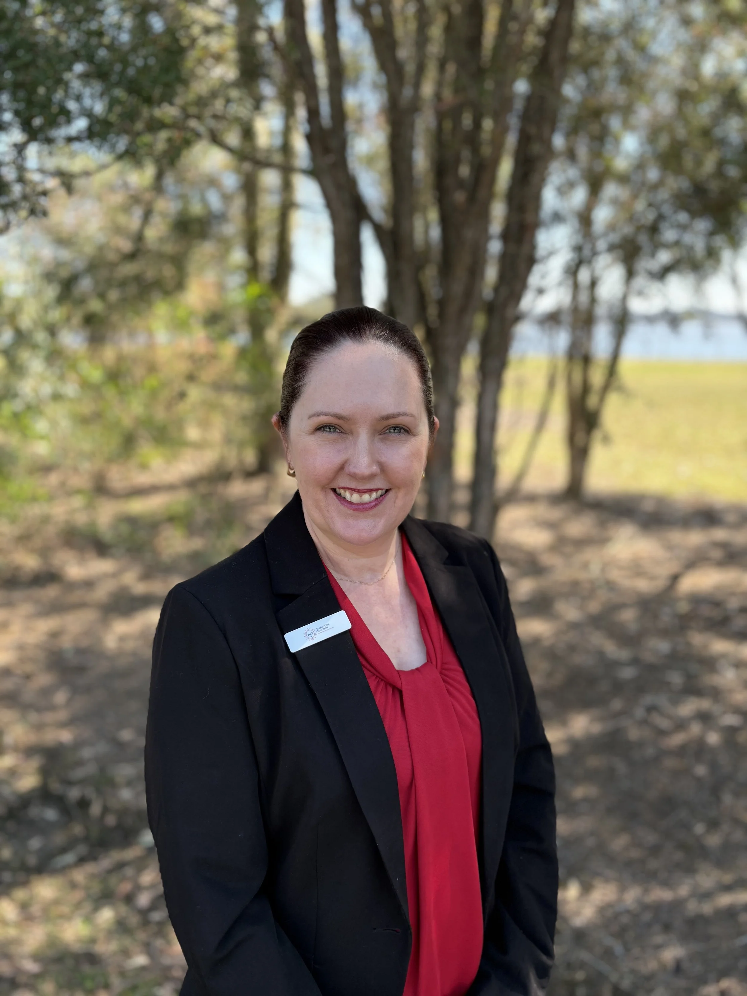 A woman smiling outdoors in front of trees, wearing a black blazer over a red blouse, with a name tag on her blazer.