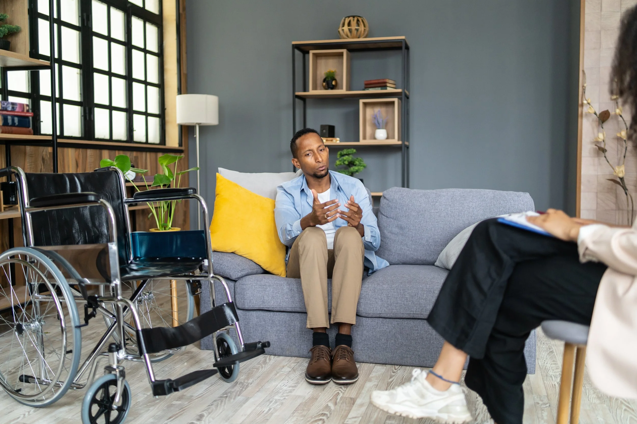 A man sitting on a gray couch in a therapy or counseling session, talking to a counselor who is taking notes. There is a wheelchair nearby, and the room has decorative shelves and a window.