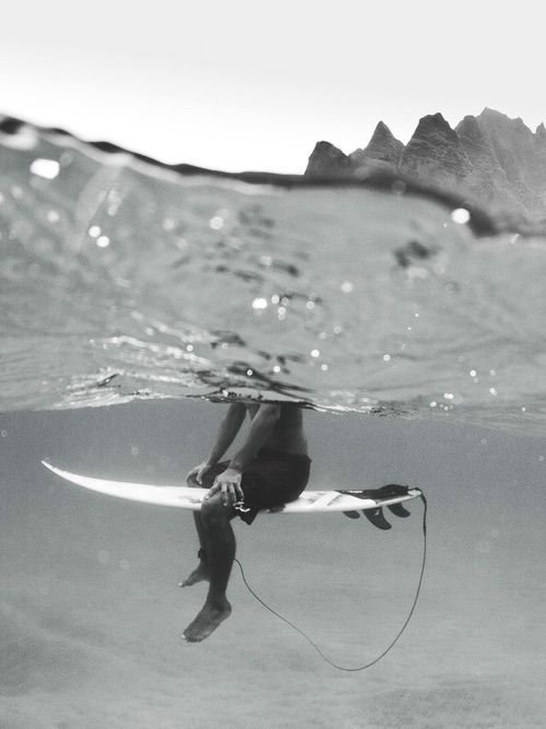 A person wearing a wetsuit and holding a surfboard, hanging upside down in the water near a shoreline with mountains in the background.