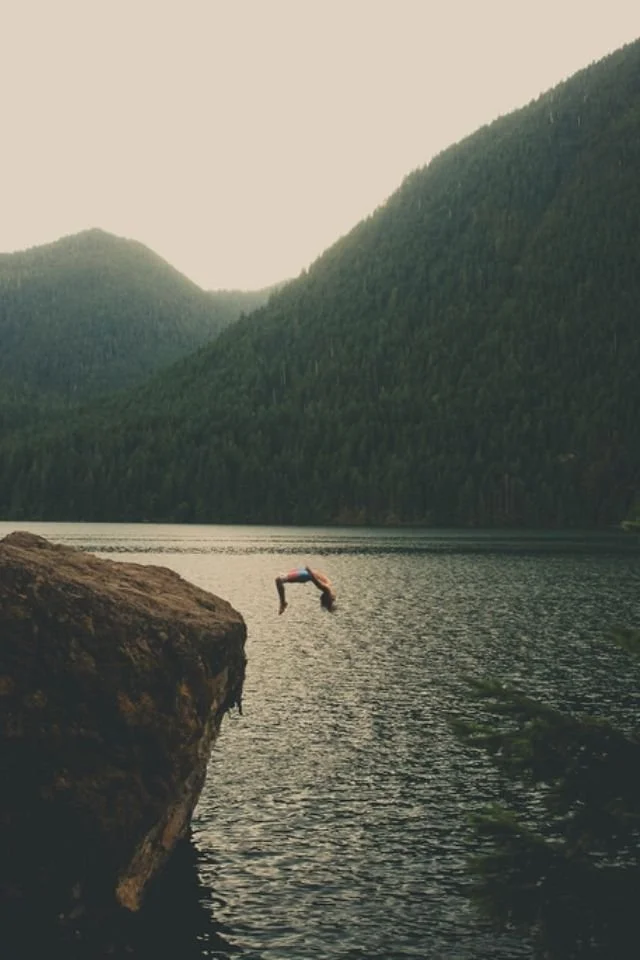 A person diving off a rocky ledge into a lake surrounded by forested mountains.