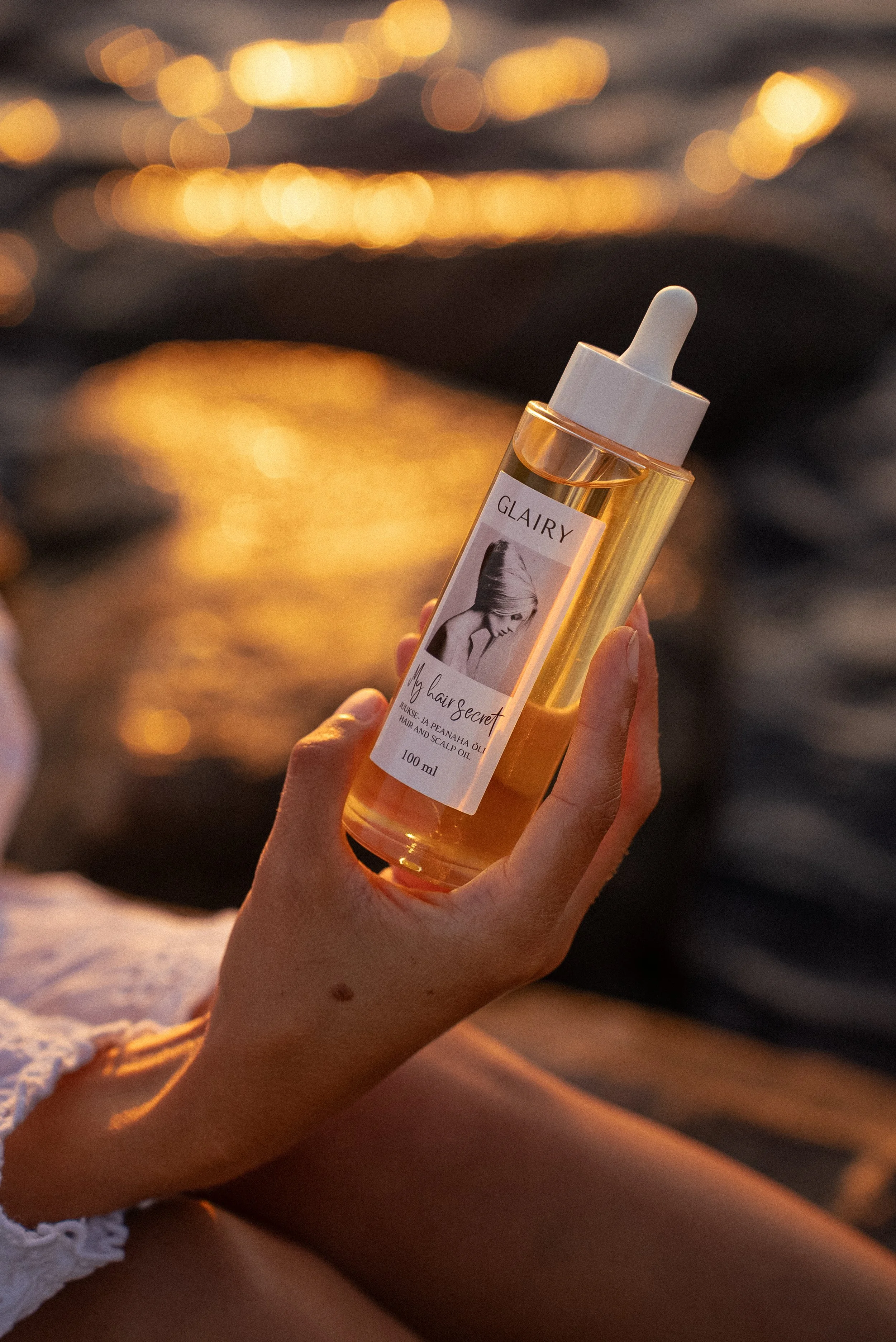 Käsi hoidmas juukseõli pudelit päikeseloojangu taustal
