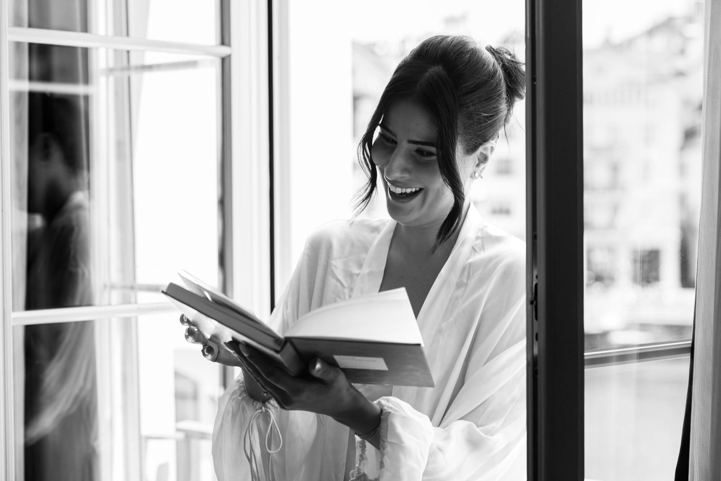 Schwarzweiss: die Braut im Morgenmantel liest lachend in einem Buch am hellen Fenster