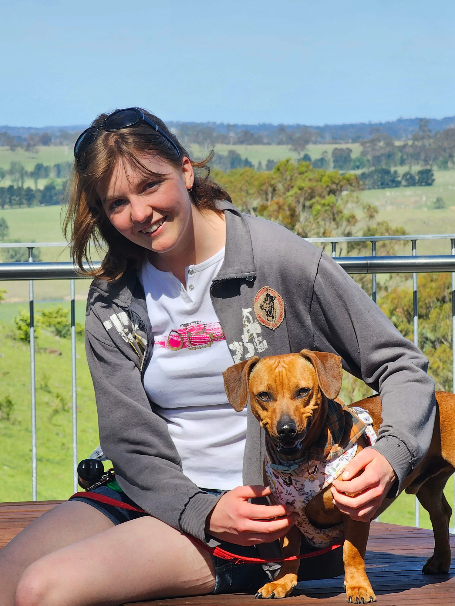 A smiling young woman (Jessica) sitting with Obi (Brown daschund) on a wooden bench at a lookout. There's green paddocks, river and trees in the background. Jess has on shorts and a white T-shirt, and Obi is wearing a colourful bandana.