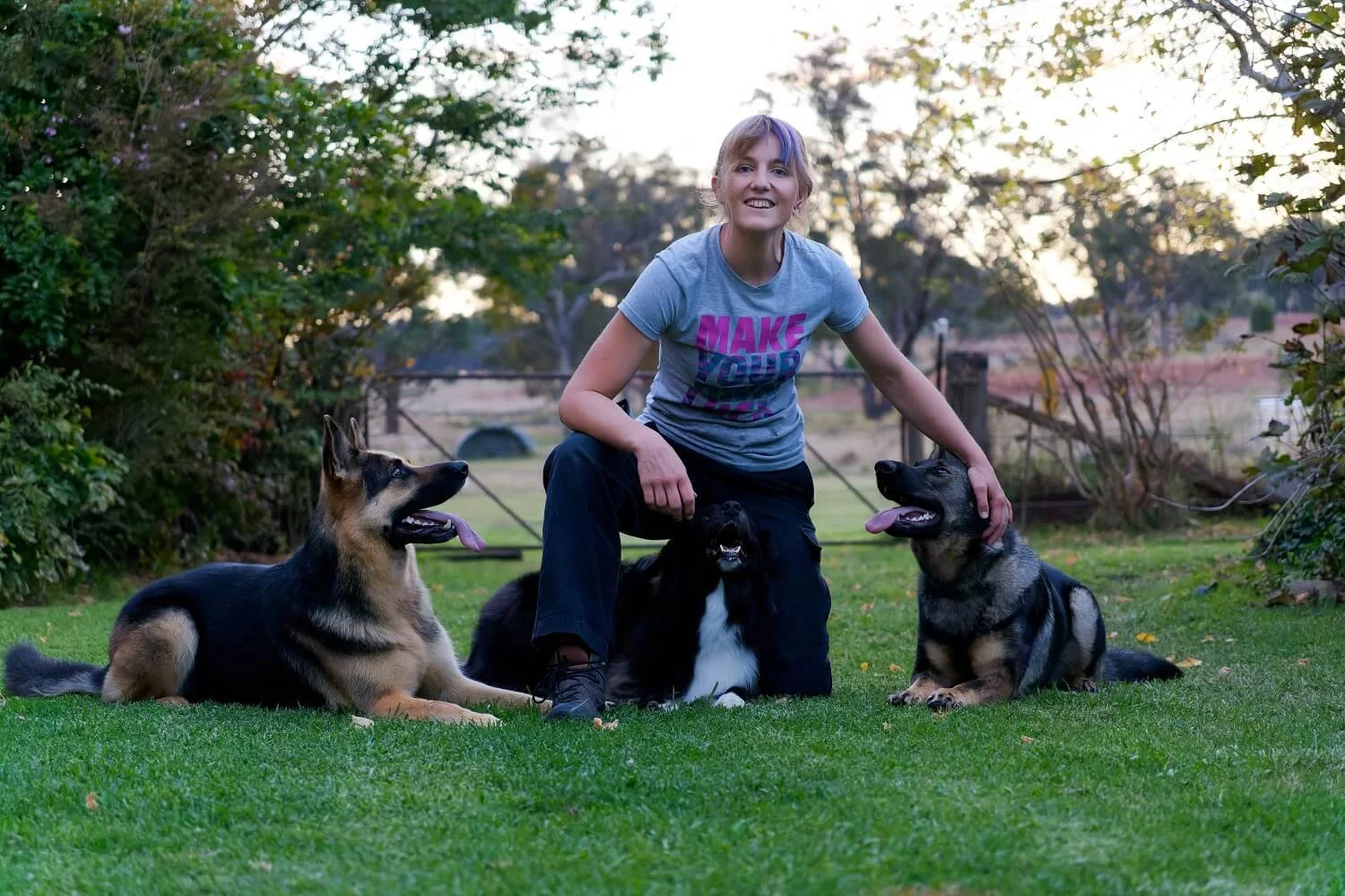 Alex kneeling on mown grass with Mischa (Black & Tan German Shepherd), Fable (Black & White Aussie Shepherd) and Addi (Sable German Shepherd) all in down positionr. Alex is patting Addi, smiling, and wearing a grey t-shirt & black cargo pants.