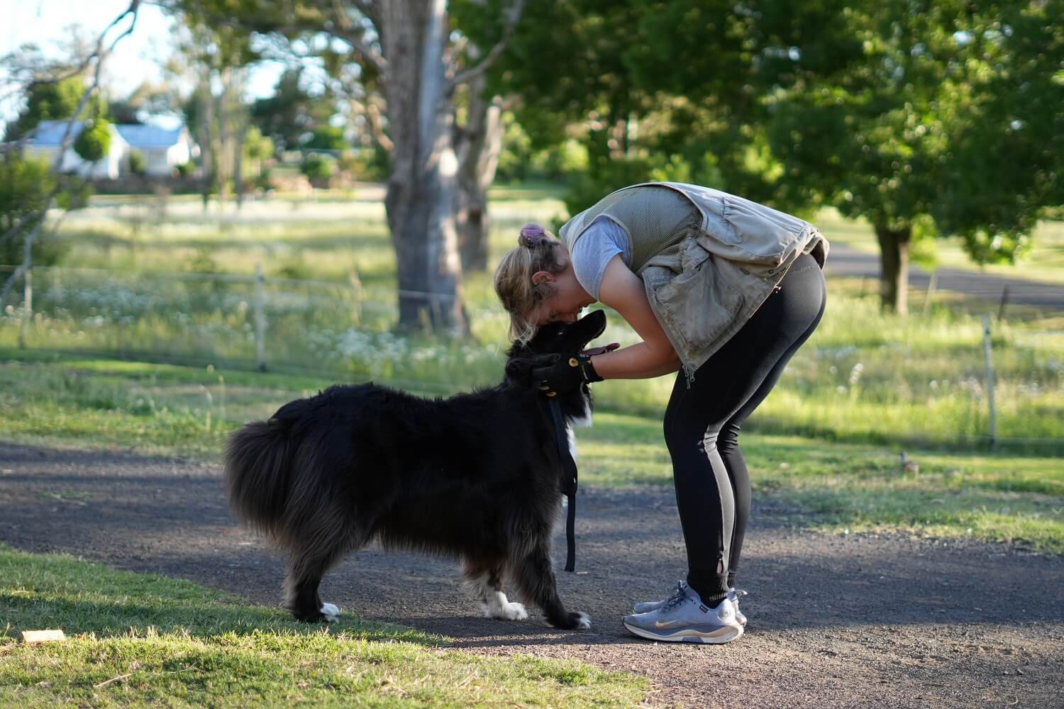 Alex and Fable (Aussie Shepherd) on a gravel path with grass and eucalypts in the background. Alex is bending down to lift Fable's face to hers in a loving, trusting gesture of companionship and love.r