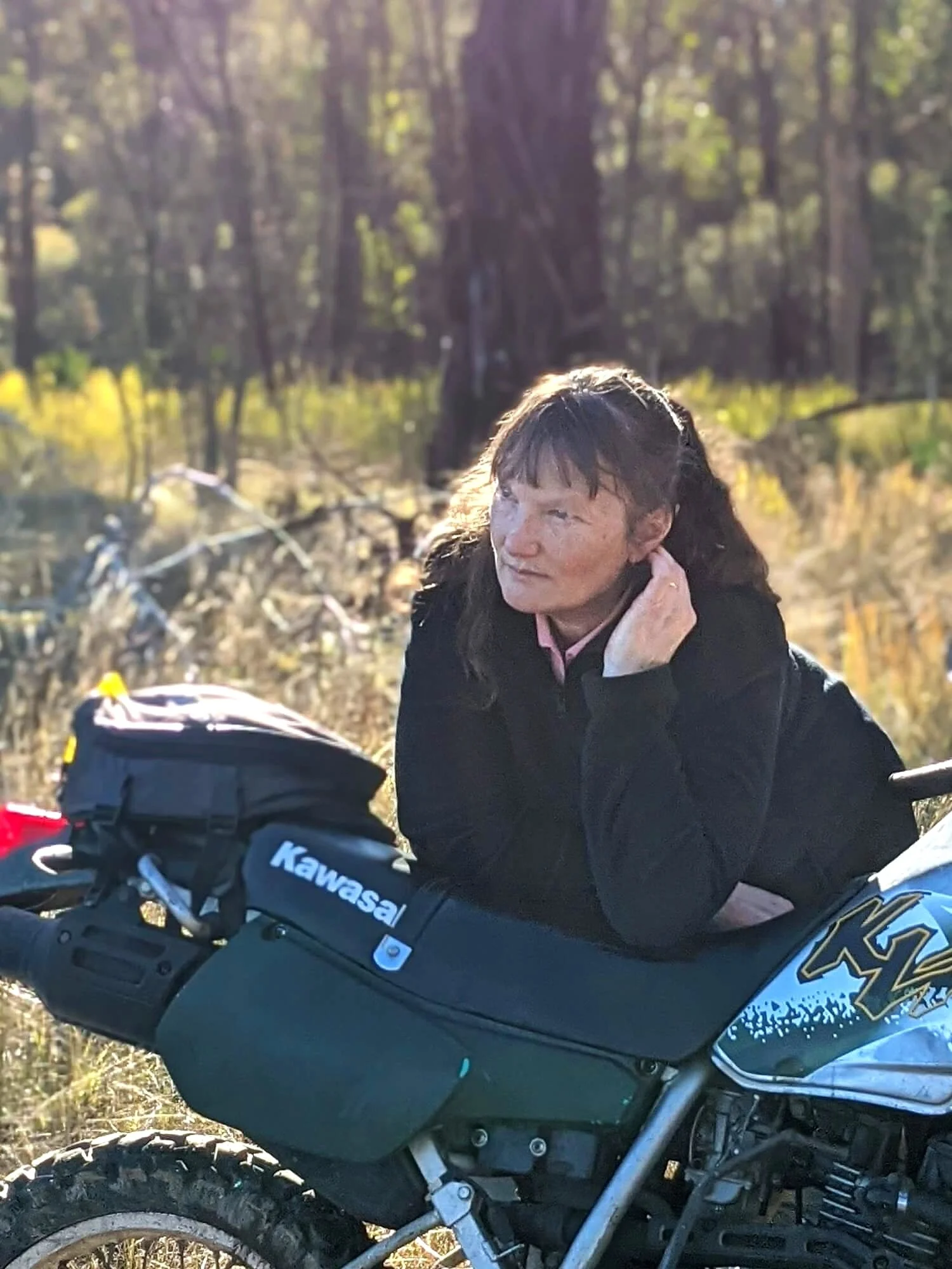 A woman with long dark hair is leaning on a Kawasaki dirt bike outdoors in a wooded area during daytime, with sunlight filtering through trees in the background.