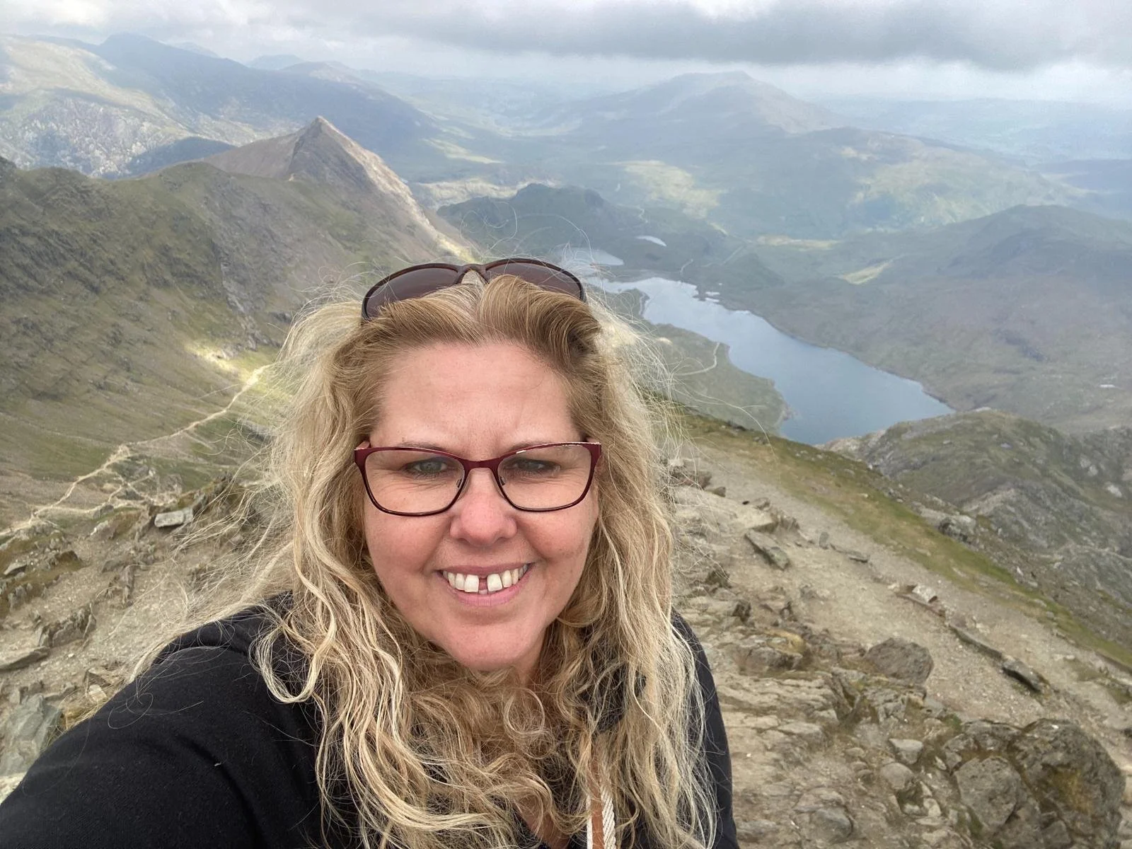 A woman taking a selfie on a mountainous trail with a lake and mountains in the background.