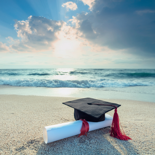 Graduation cap and diploma paper on the beach with ocean waves and a cloudy sky during sunset.