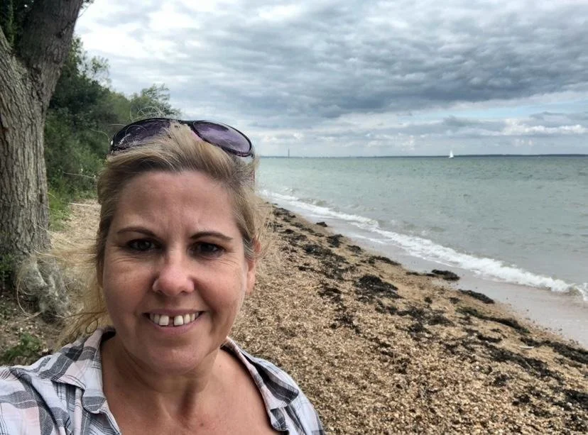 A woman taking a selfie on a beach with cloudy sky, trees, and ocean in the background.