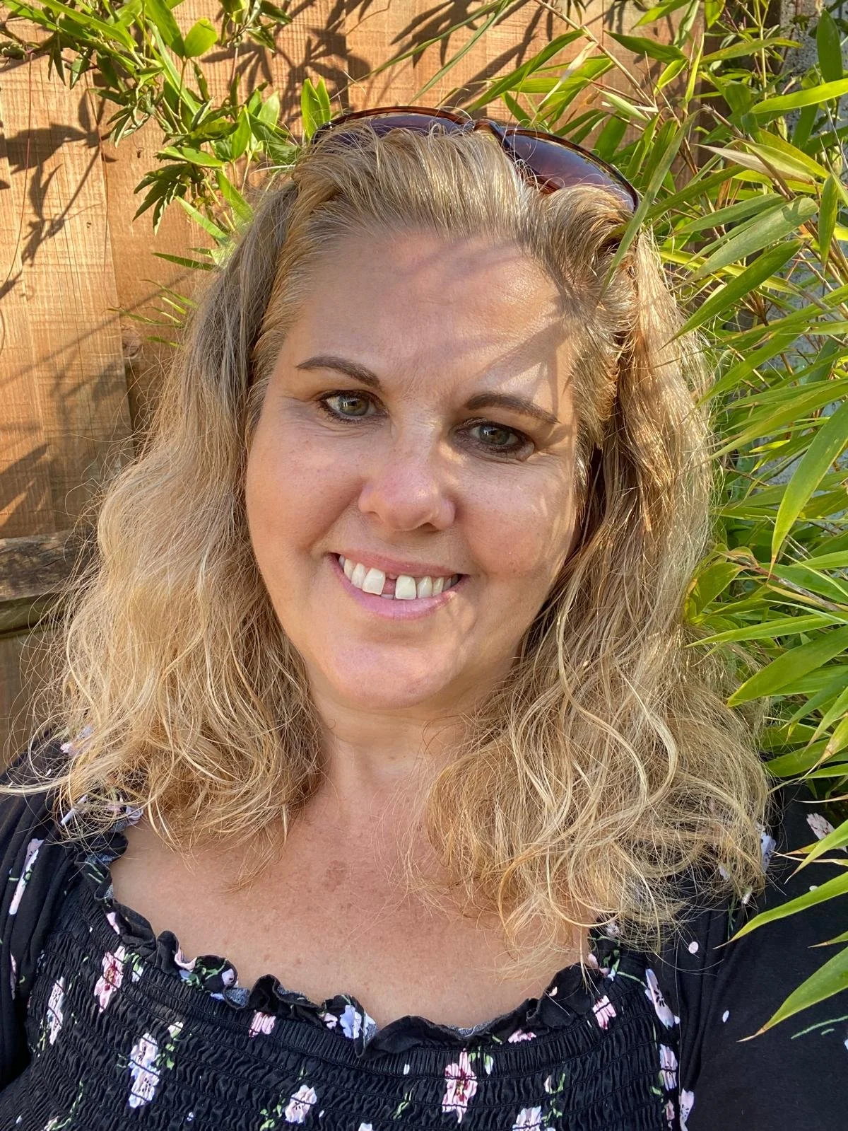 A woman with blonde, curly hair is smiling in an outdoor setting with green plants and a wooden fence in the background.