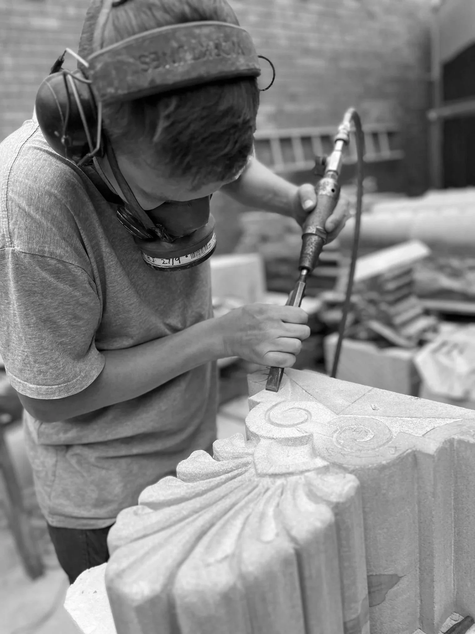 A person wearing a hard hat and safety mask is using a rotary tool to carve a decorative pattern into a piece of wood with swirling designs. The background shows a brick wall and workshop tools.
