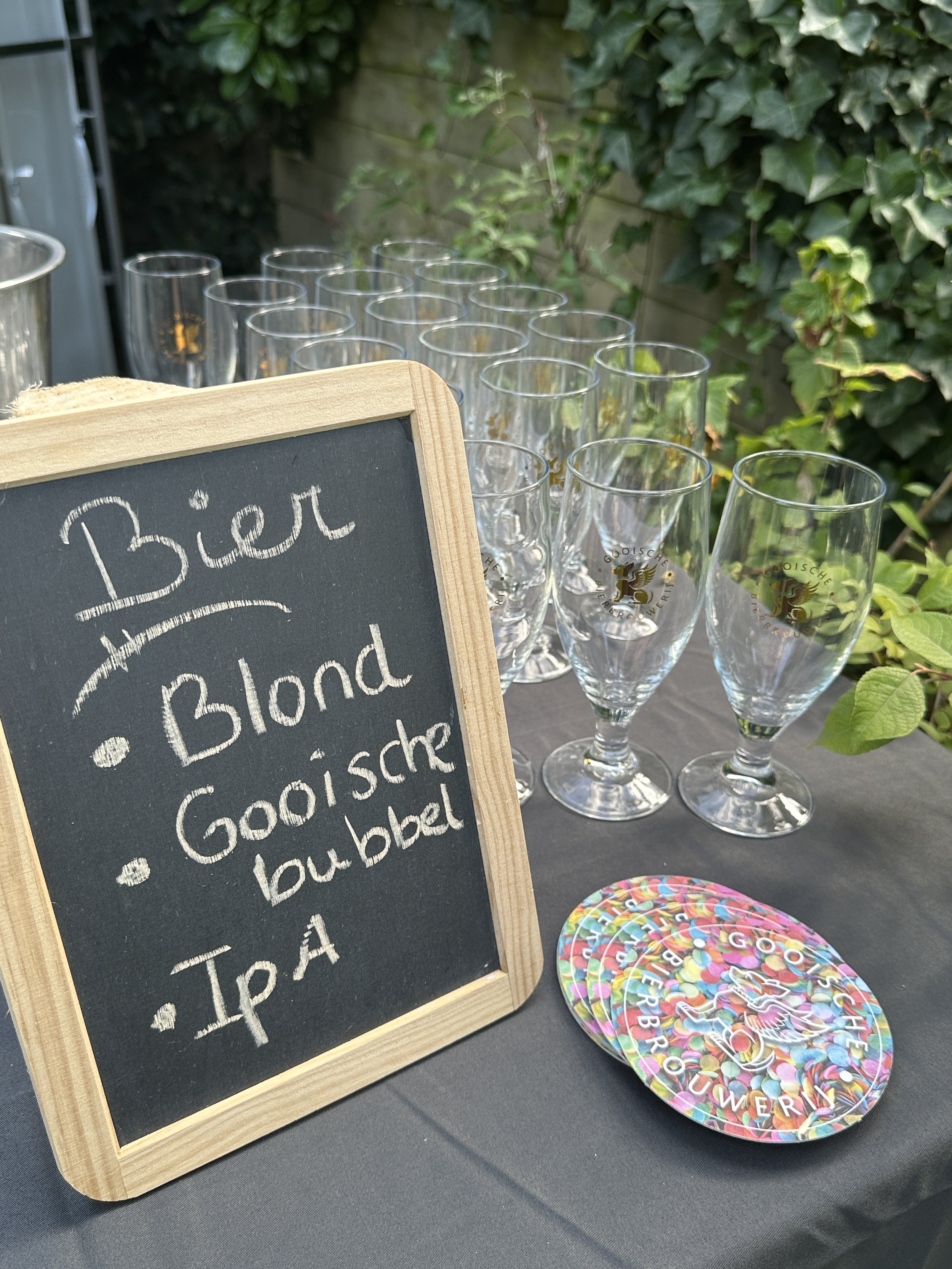 Een tafel met glazen en een klein schoolbord met de tekst 'Bier, Blond, Gooisiche bubbel, IPA'.