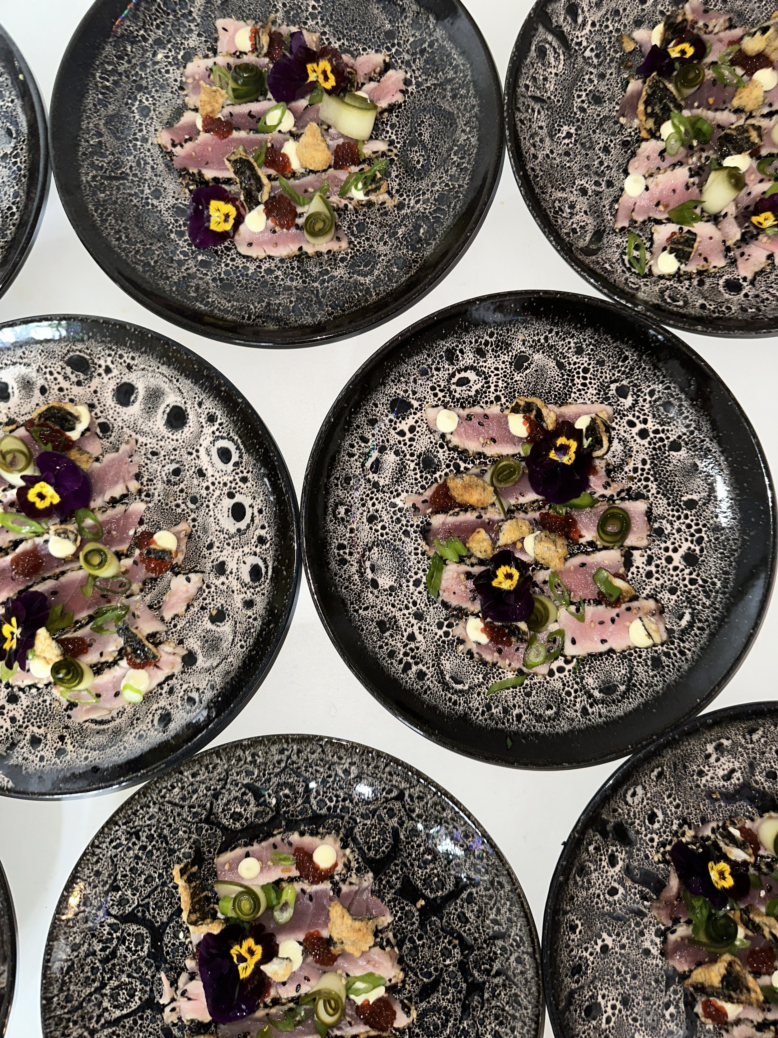 Pollepel met rauwe vis, garnituur van bloemen, groene uien, knapperige stukjes, en saus op zwarte bordjes.