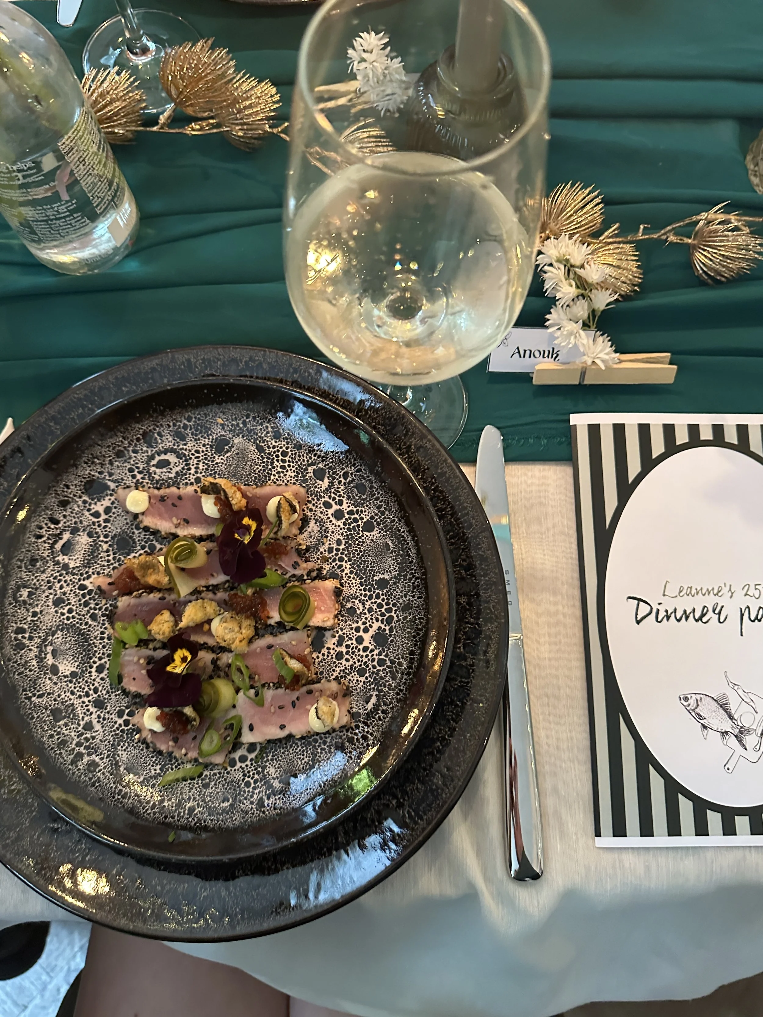 Gerecht met dunne plakjes rauwe vis, gegarneerd met groene uien, bloemen en crumpets, geserveerd op een zwart bord met een bolvormige rand, naast een glas witte wijn en een fles water op een groene tafelkleed.