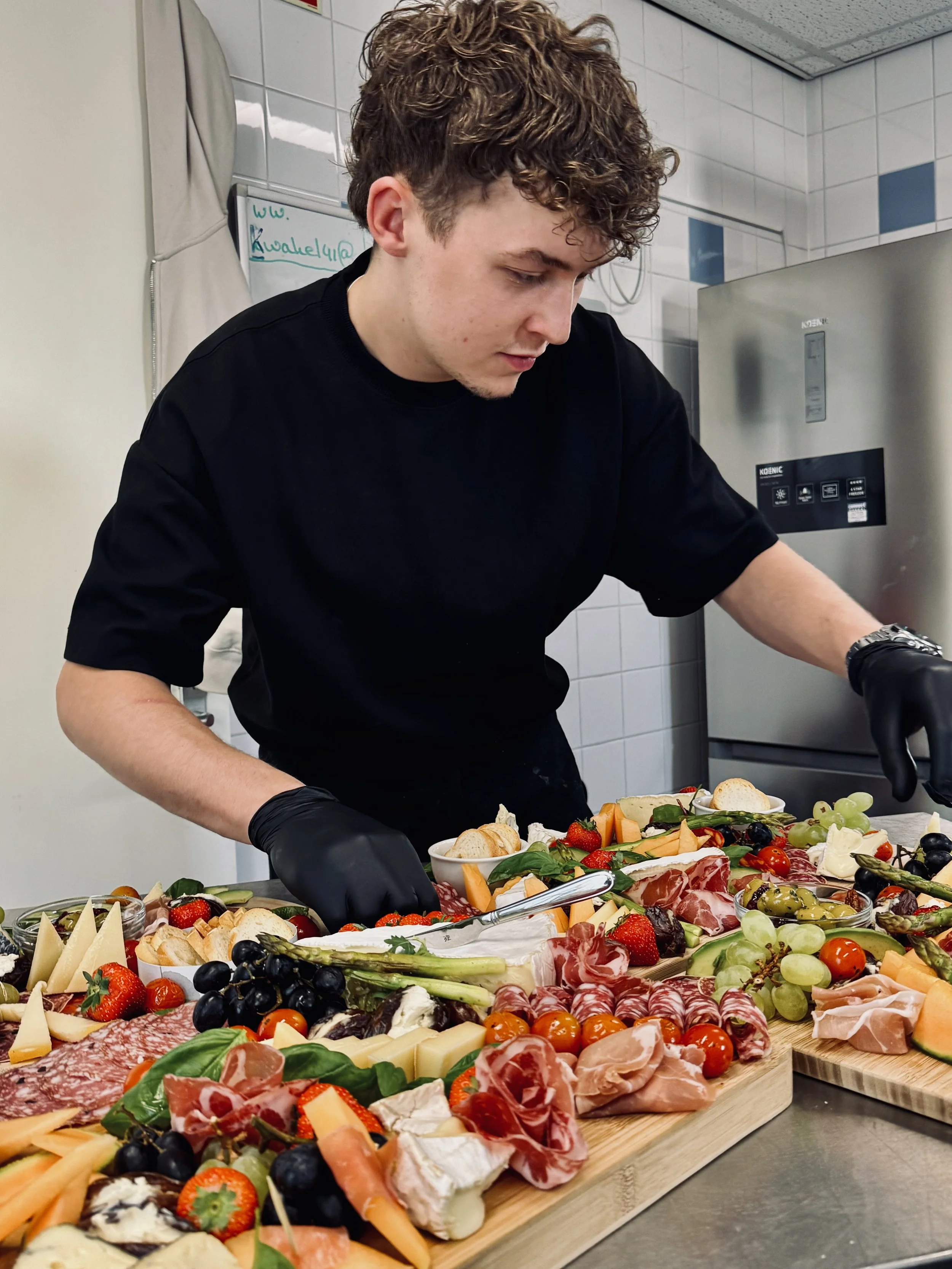 Man die een uitgebreide en kleurrijke kaas- en charcuterieplank samenstelt met diverse kazen, vleeswaren, druiven, aardbeien, tomaten, en crackers in een keuken.