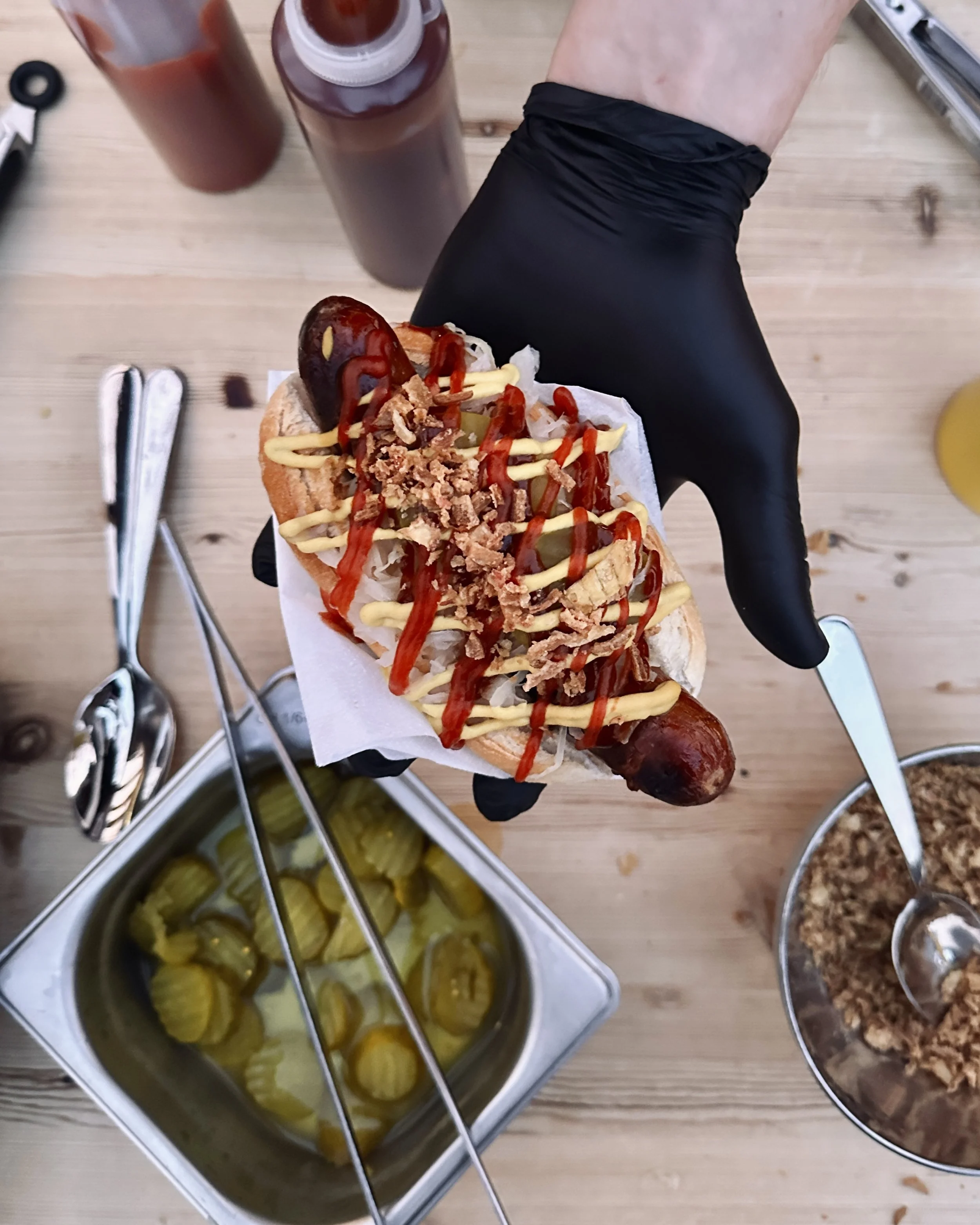 Persoon houdt een hotdog met ketchup, mosterd, geraspte kaas en uitjes. Op tafel staan potten met saus, een schaal met augurken en een kom met gehakt.