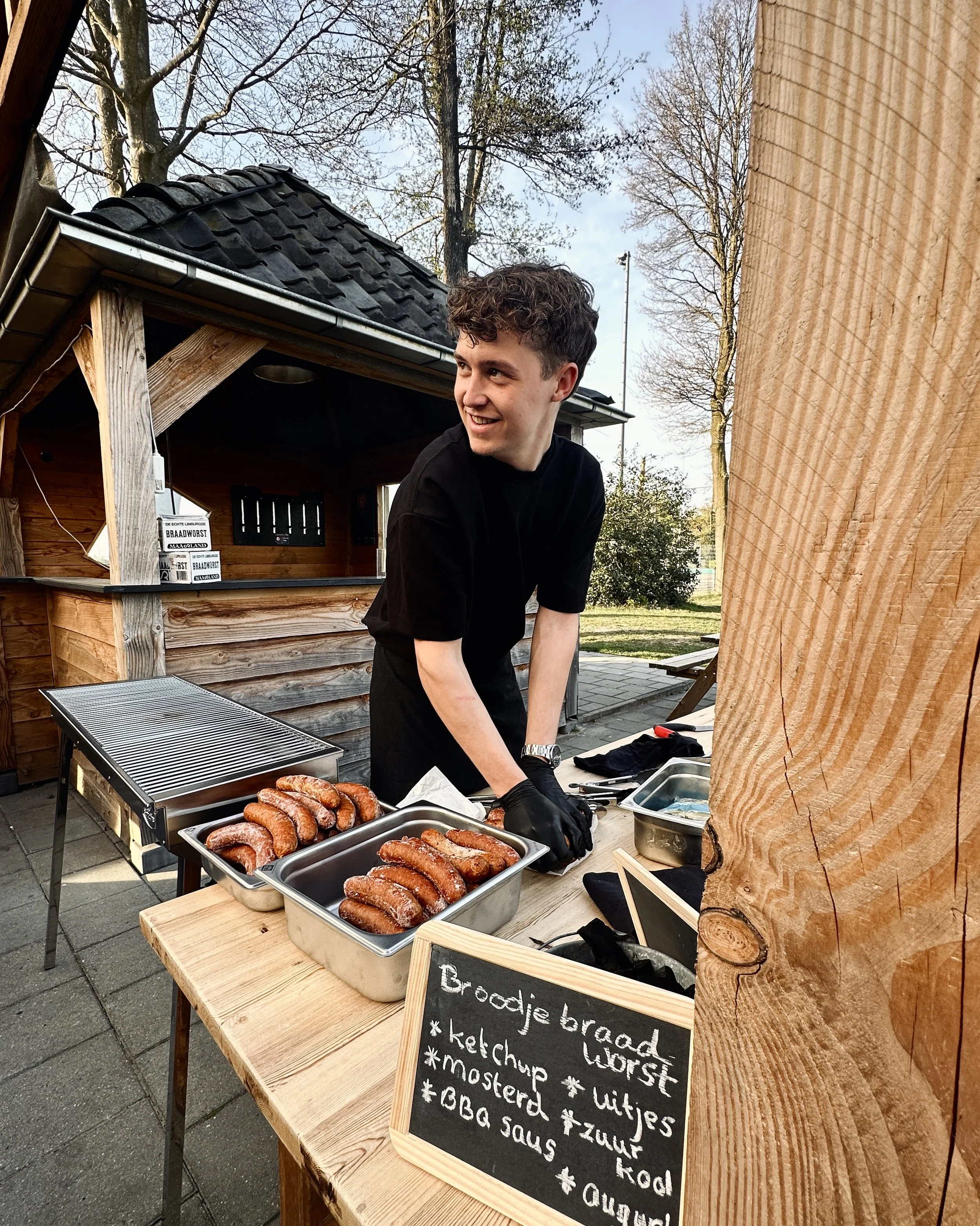 Een jonge man die op een buitenbarbecue stand staat, die worstjes klaar maakt, met een bord en een houtbordje met barbecue-gerelateerde kruiden en sauzen.