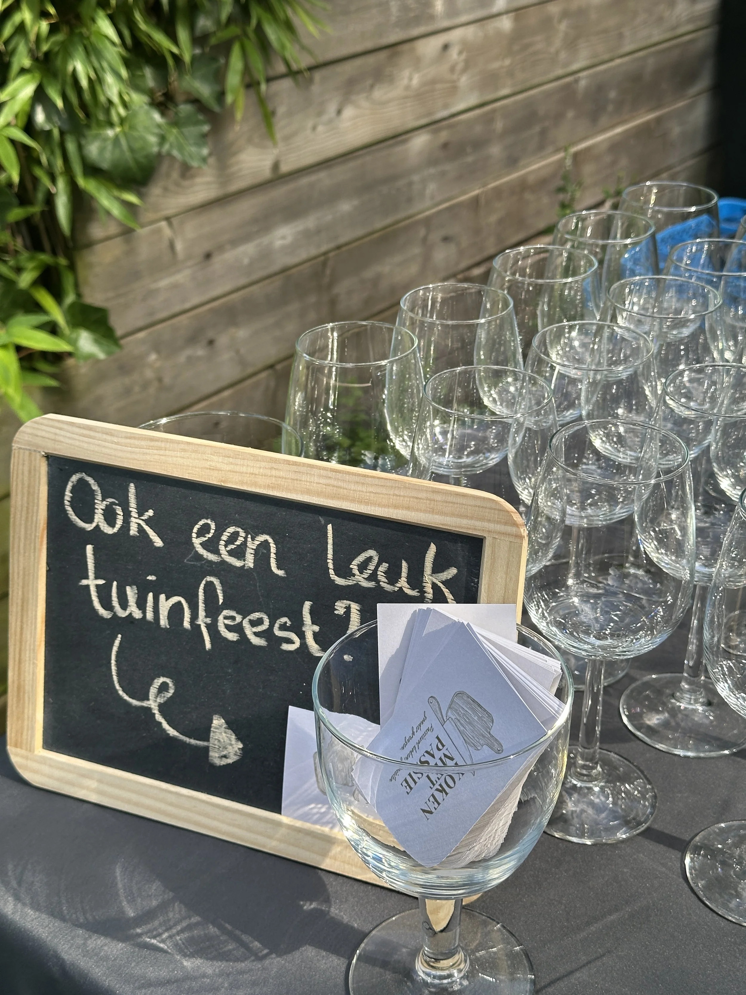 Een tafel met lege wijnglazen, een klein krijtbord met de tekst 'Ook een leuk tuinfeest?....', en een glazen kom met papieren wachtwoorden of flyers.