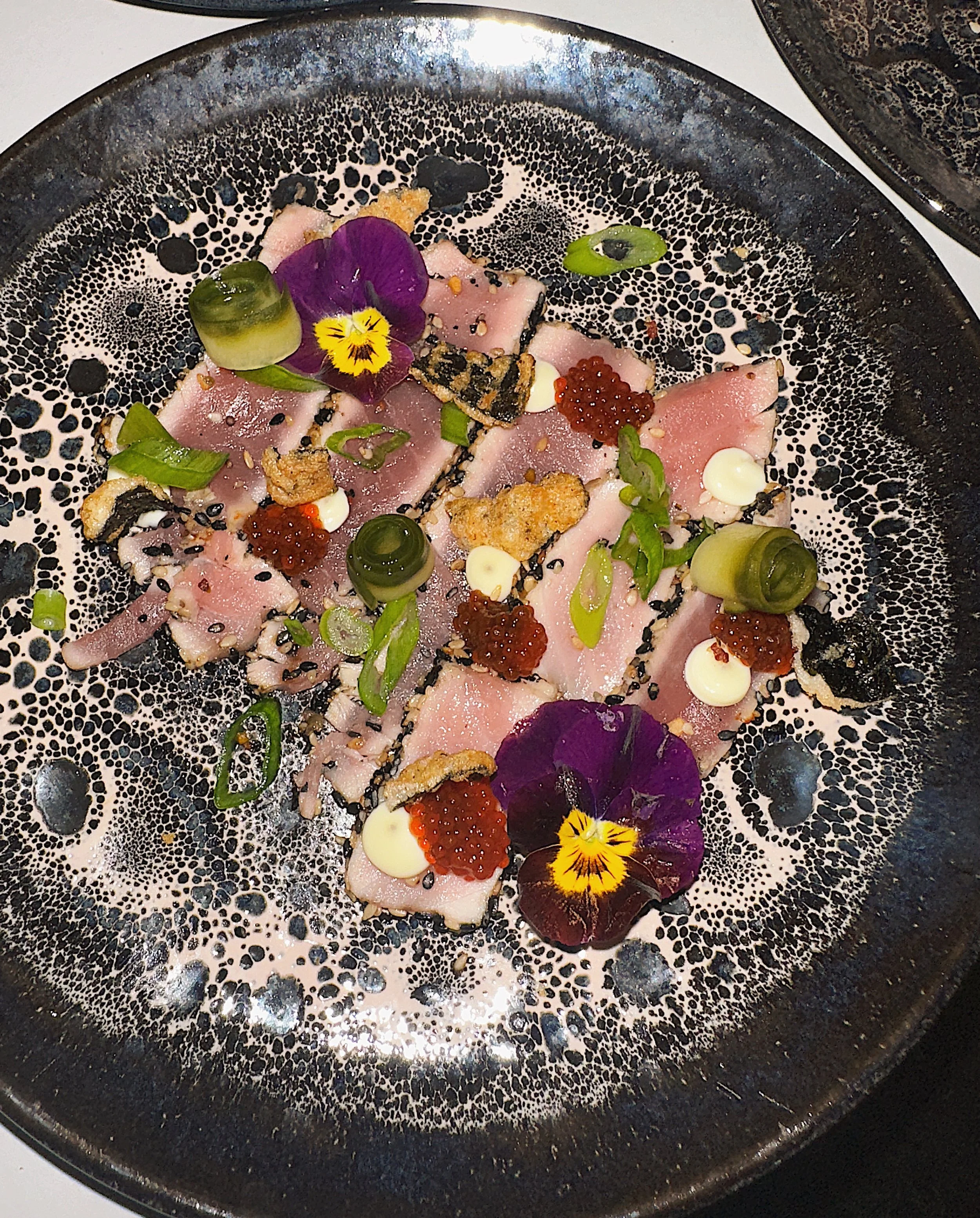 Gerecte sashimi met groene ui, rode kaviaar, zwarte sesam, eetbare bloemen, en saus op een decoratief zwart bord.