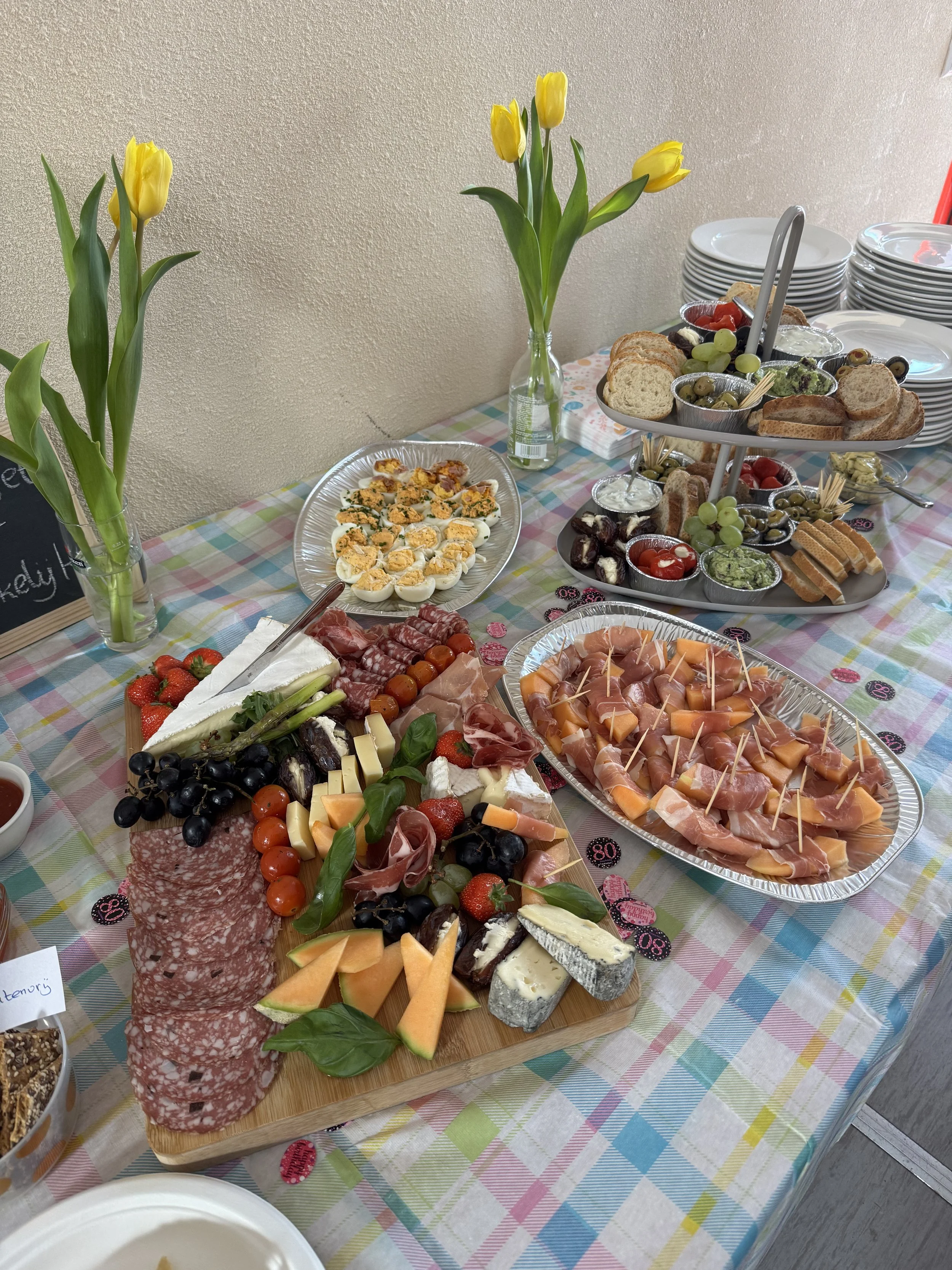 Een uitgebreide borrelplank met verschillende kazen, vleeswaren, groenten, druiven, aardbeien, en crackers, met twee vazen met gele tulpen op een tafel met een kleurrijk tafelkleed. Daarnaast een twee verdieping schaal met diverse broodjes, dips, en 