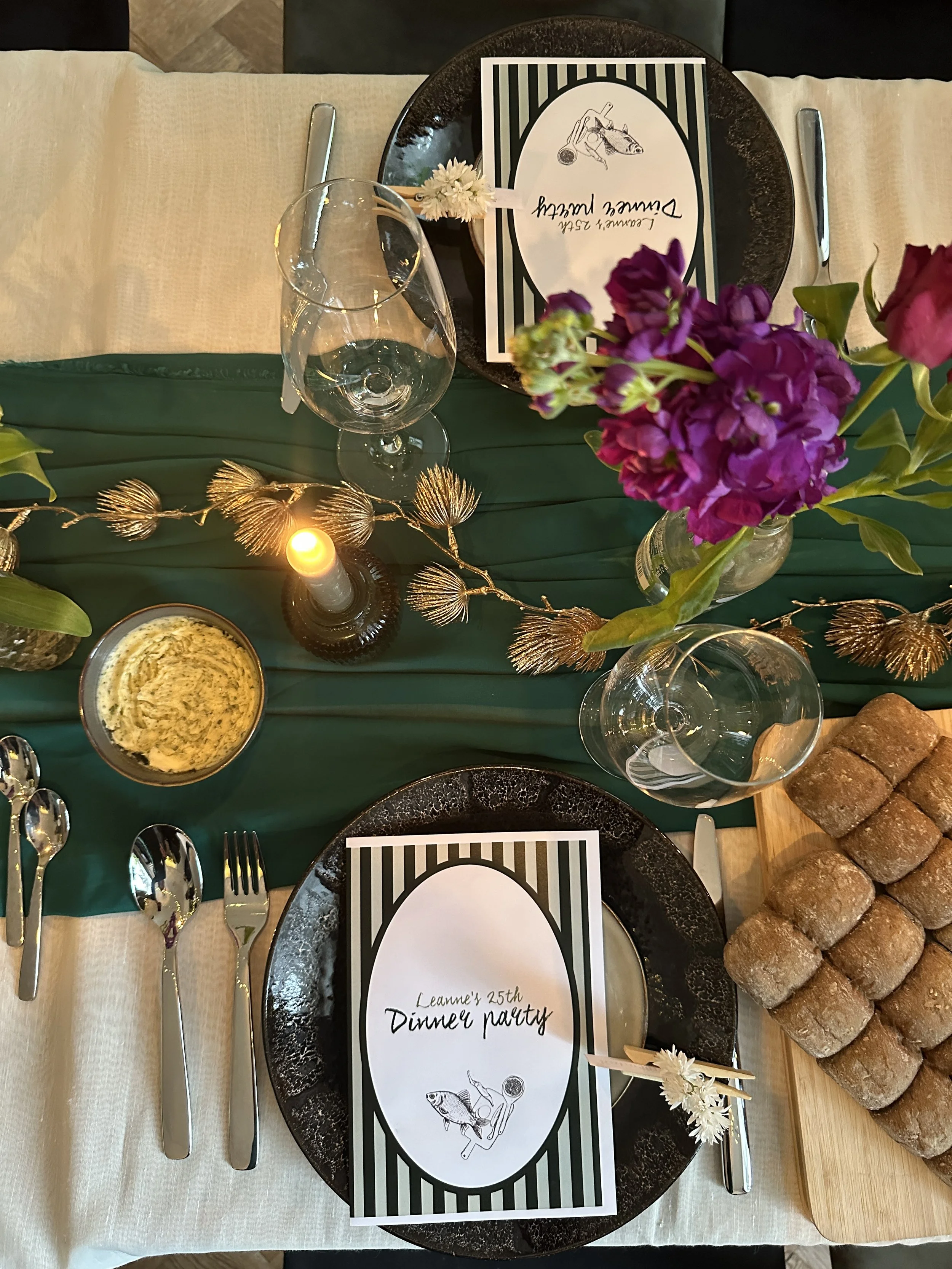 Tafel gedekt voor een etentje met kaarten die 'Leanne's 25th Dinner Party' aangeven, bloemen, glazen en een broodplank.