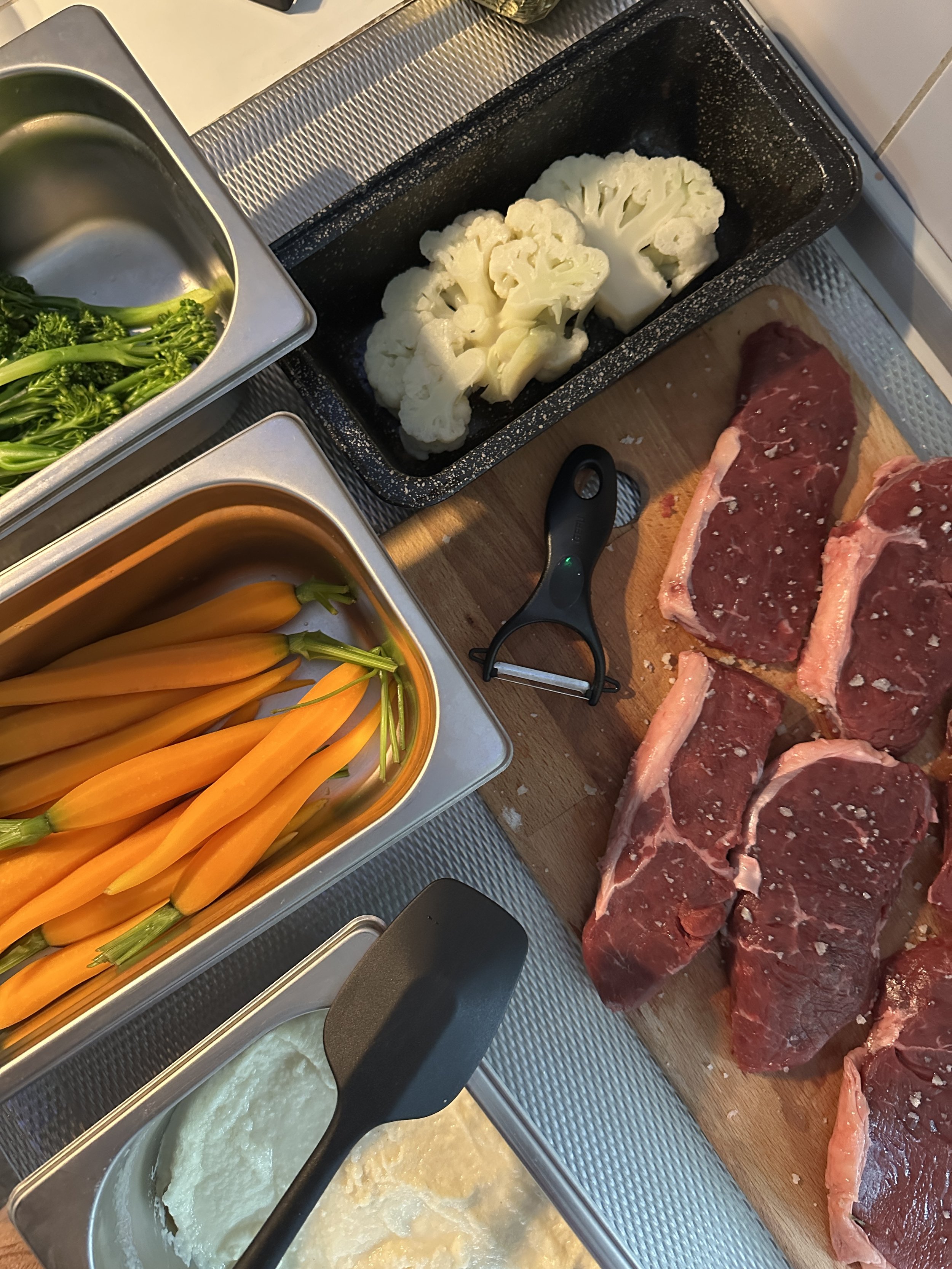 Verschillende ingrediënten voor een gerecht: broccoli, gele wortelen, rundvlees steaks, bloemkool, en aardappelpuree.