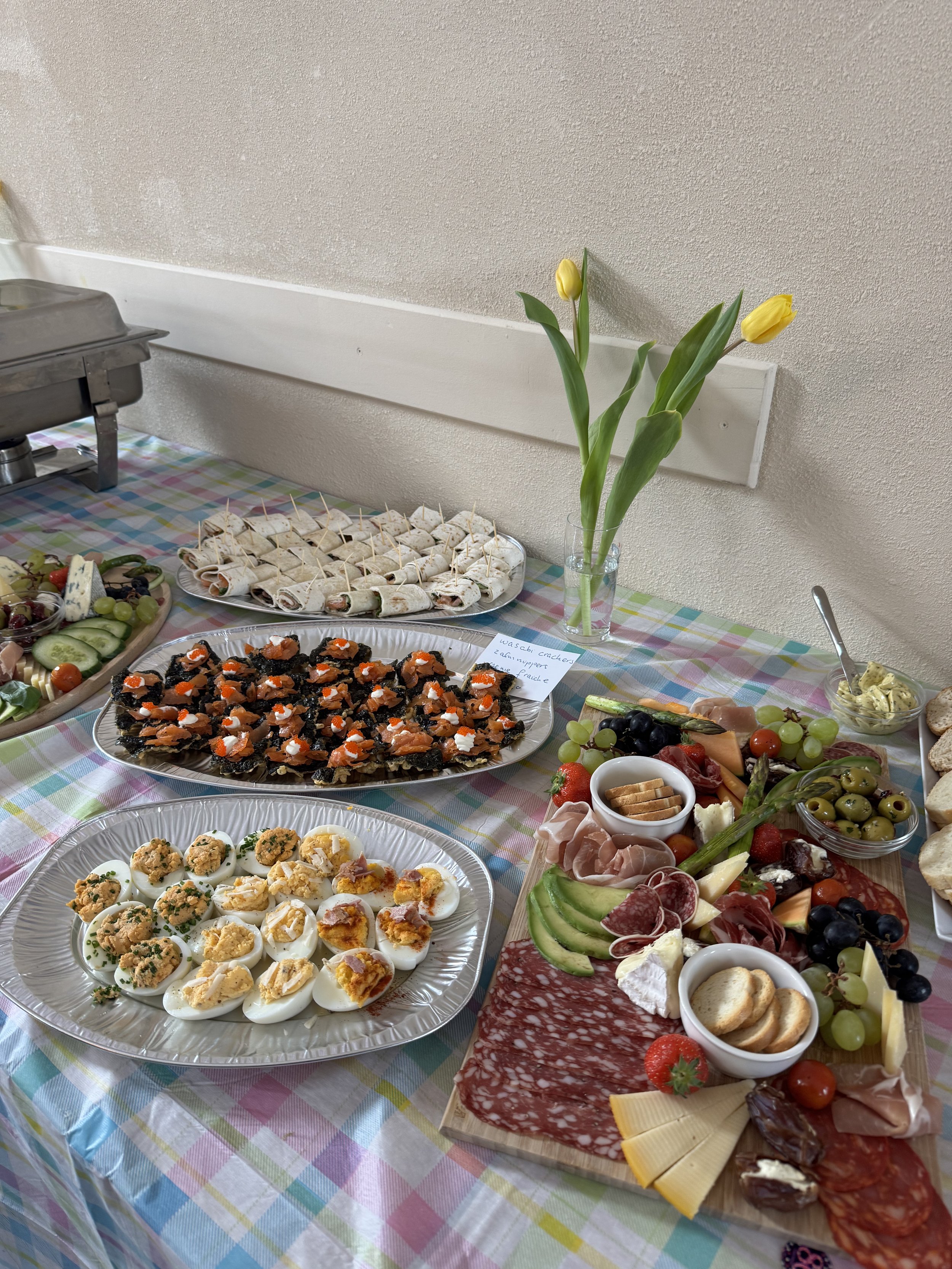 Een tafel met diverse hapjes en snacks, waaronder gekookte eitjes, chips, vleeswaren, kaas, druiven, aardbeien, en een vaas met gele tulpen.