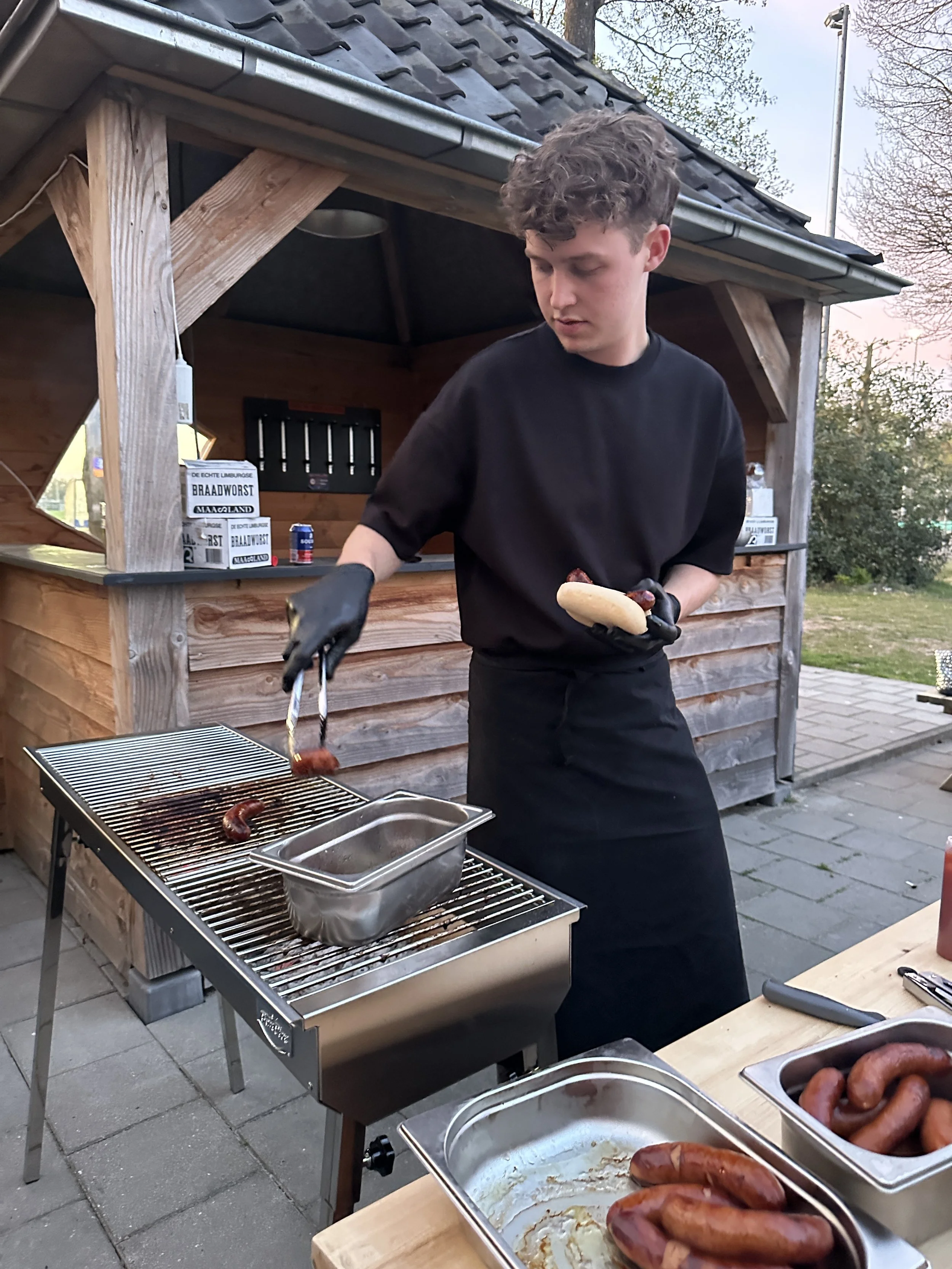 Jongen die worstjes op de grill bereidt in een buitenkeuken, met een broodje in de hand, terwijl het buiten donker wordt.