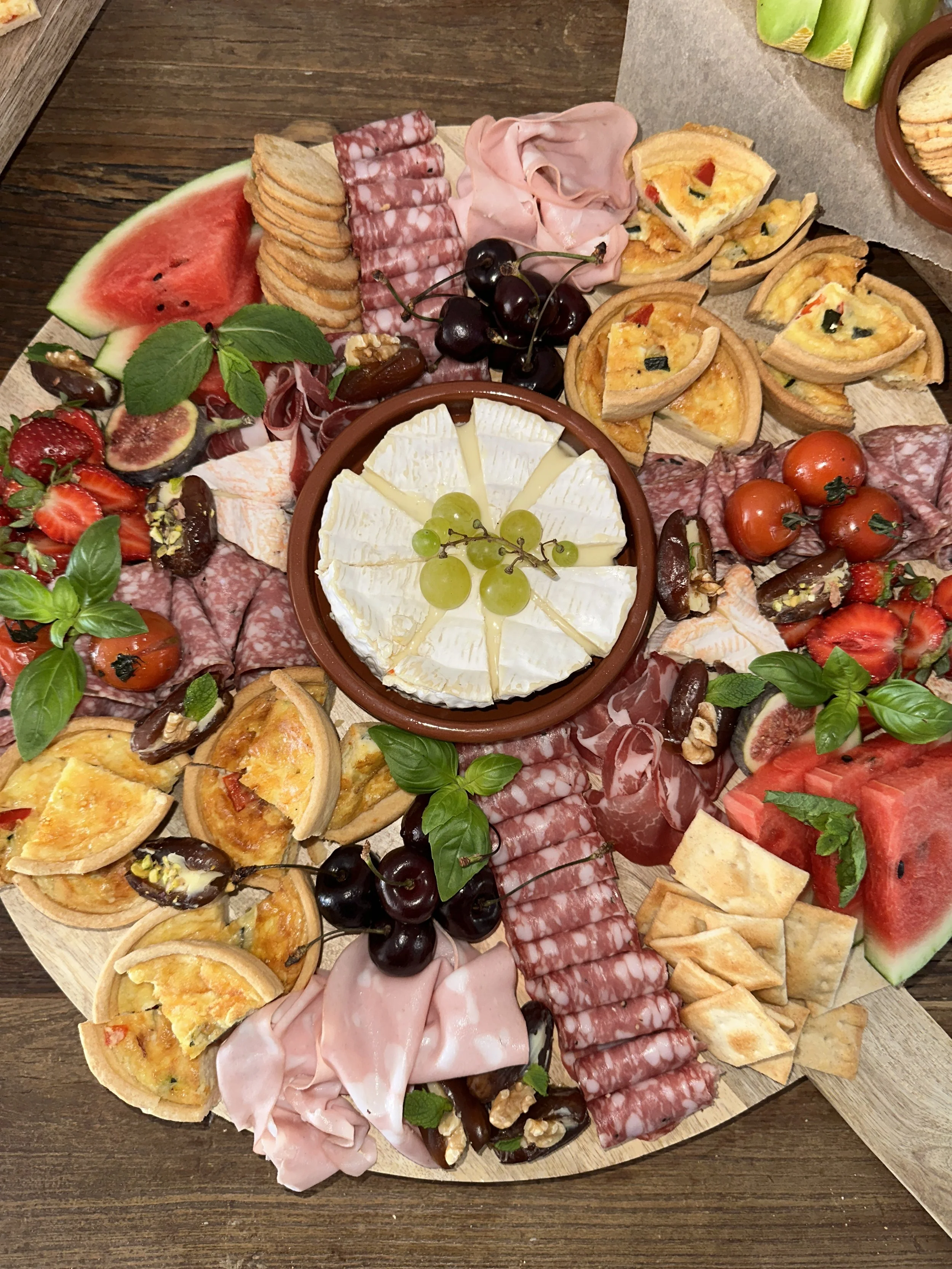 Een grote ronde antipasti-plank met diverse kazen, vleeswaren, vers fruit, kleine taartjes, tomaten, druiven, aardbeien en verse kruiden zoals basilicum.