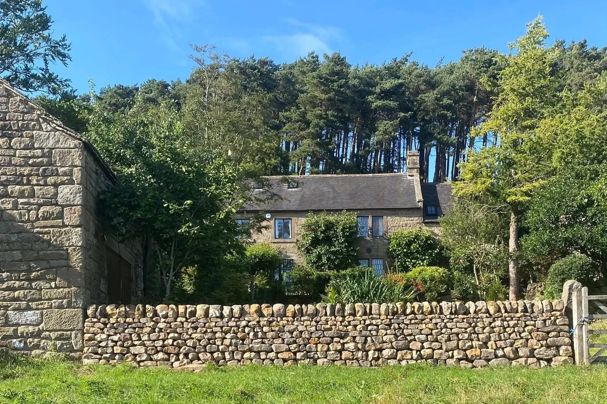 Coursed dry Stone wall house behind a stone wall with trees and greenery in the front yard, clear blue sky above. Belper, Longstone, Youlgrave, Cladding, Dry stone effect