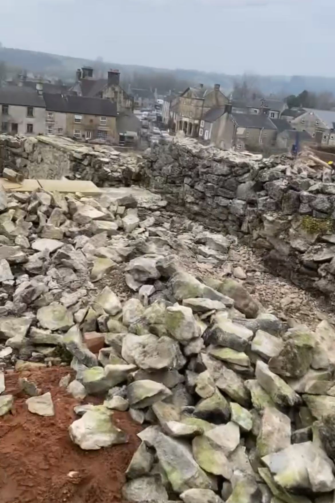 Ruins of a dry stone wall and scattered rocks in a construction or demolition site with a small town and hills in the background. Dry stone waller near me. Bakewell. Stonework. Chapel en le frith. Stockport, Manchester. Hayfield