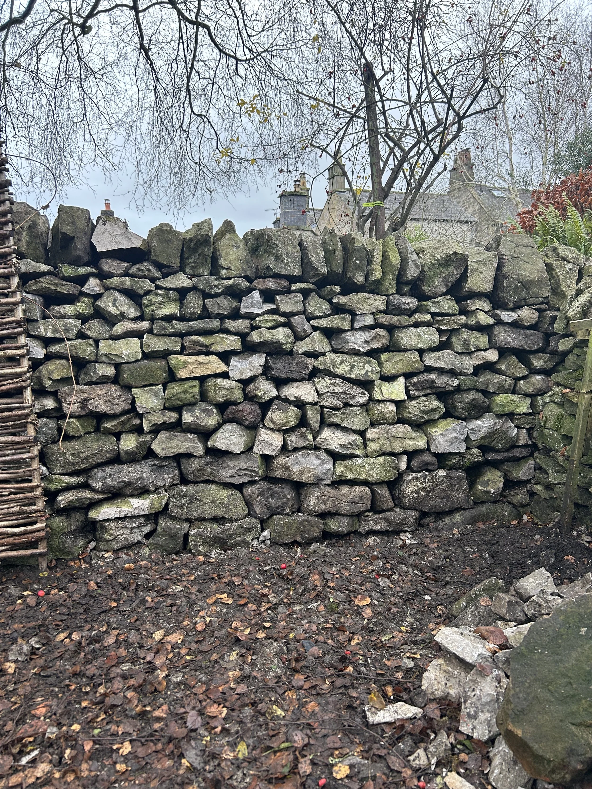 A stone wall made of irregularly shaped rocks stacked on top of each other, with a rustic wooden fence on the left and fallen leaves and small rocks in the foreground. Trees with bare branches are visible at the top, and houses are seen in the backgr