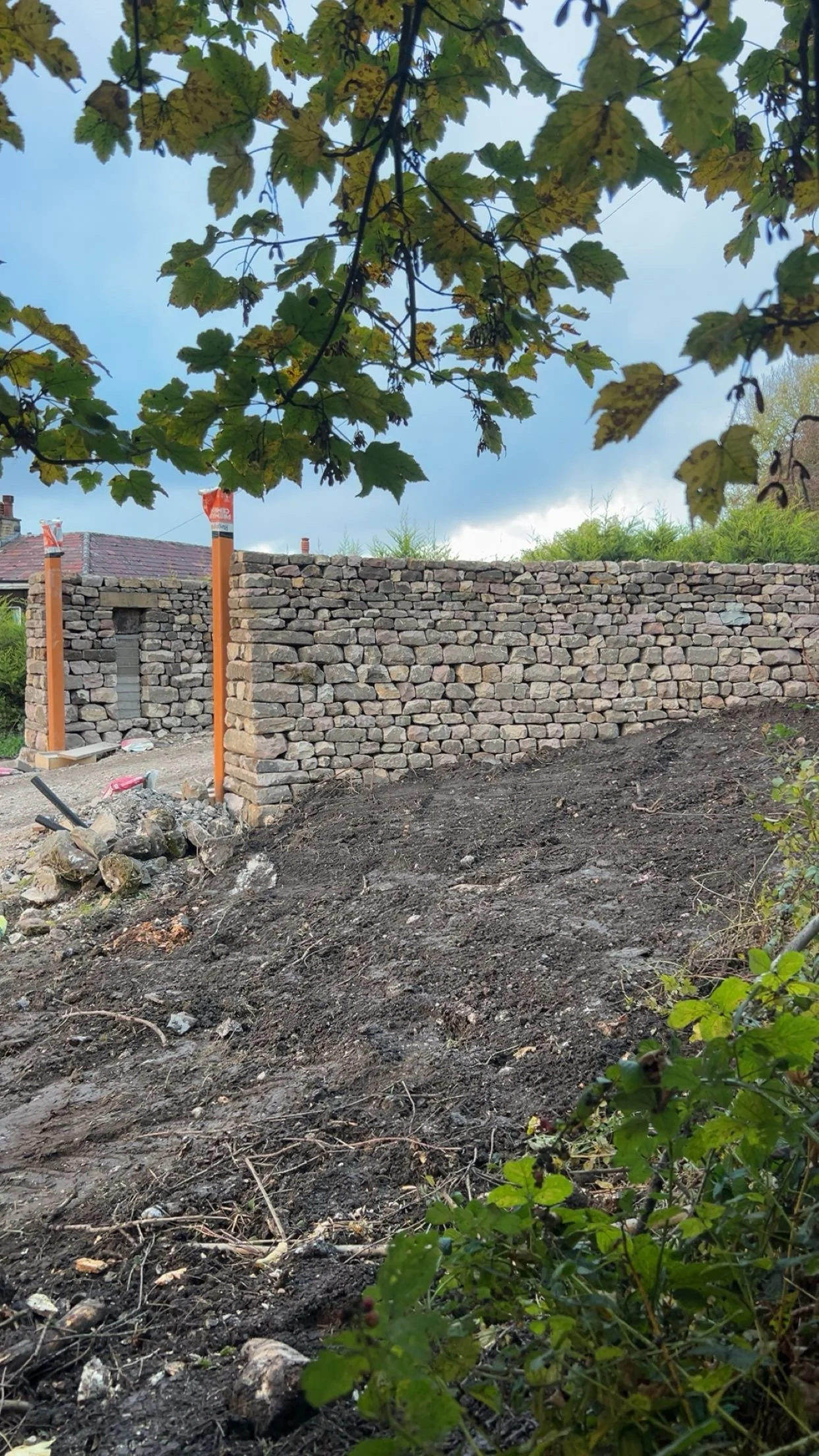 A stone wall under construction with orange wooden posts and construction tools on a dirt ground, with overhanging green leaves and a cloudy sky in the background. eNTRANCE, DRY STONE PILLARS, ENTRANCE pillars, gateway, gateposts, stonework