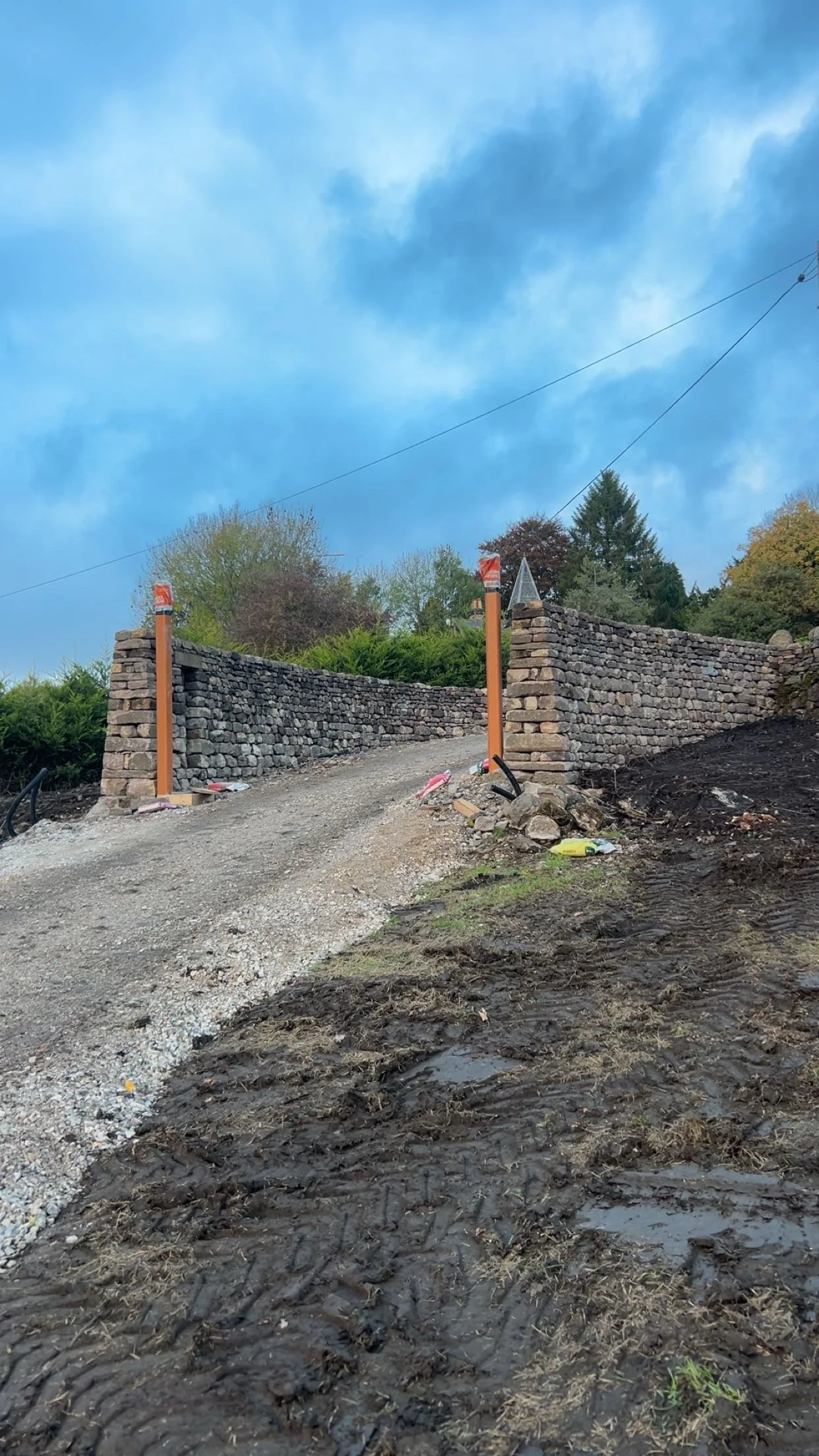 A construction site of a stone wall with orange poles at the entrance, dirt and gravel path, and trees in the background under a blue sky.stonework, stonemason, dry stone walling association, cscs card, cladding, retaining, block work, Cheshire, gard
