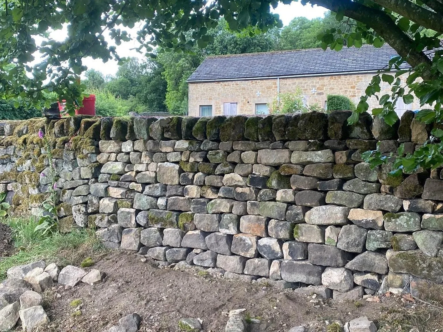 A stone wall made of irregularly shaped rocks, with some moss growing on the top stones, in front of a house with a slate roof and light-colored brick walls, surrounded by greenery. Garden dry stone wall rebuild repair emergency derbyshire near us
